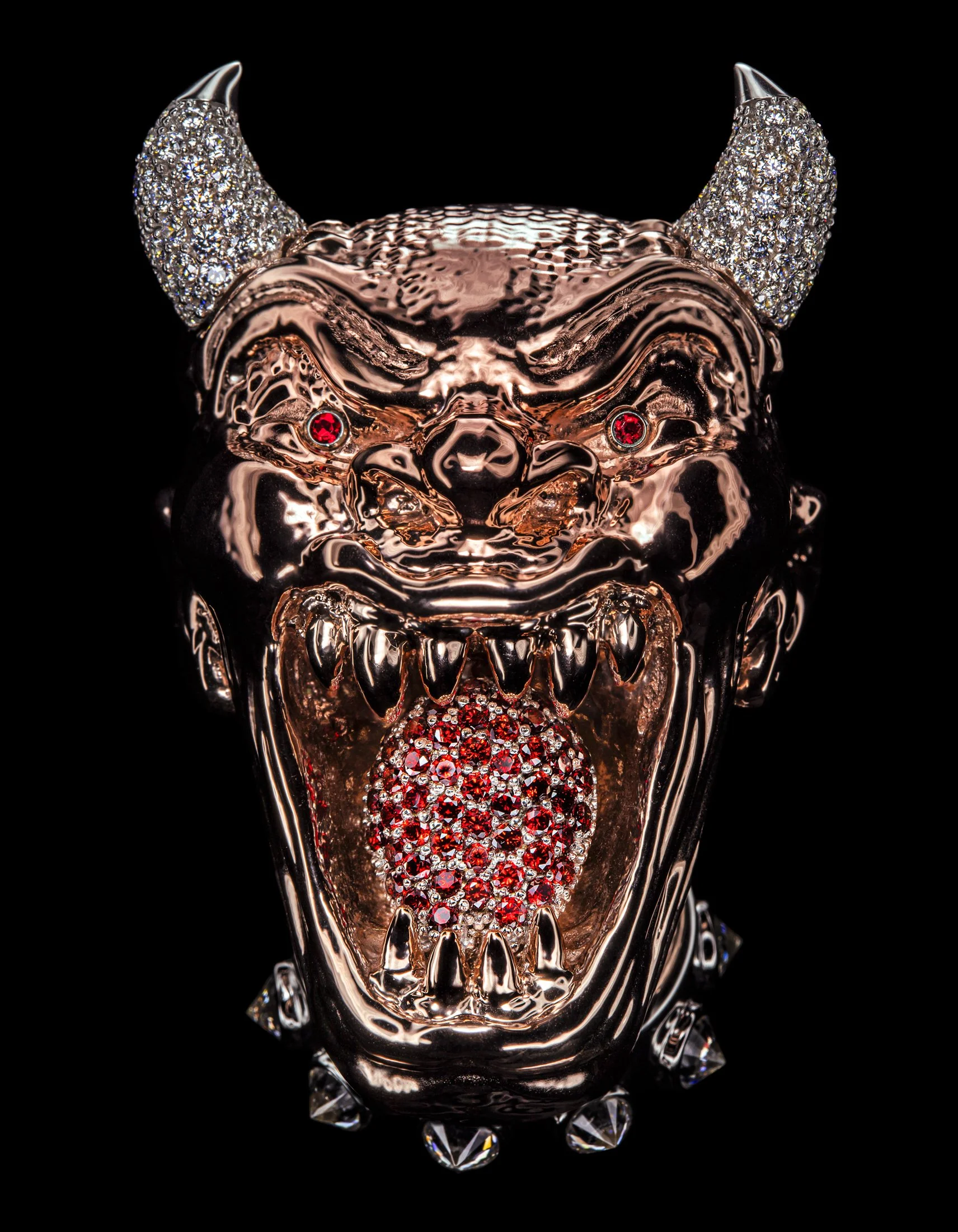 A metallic bull head ornament with ruby eyes, adorned with rhinestones on the horns, featuring a mouth open with a diamond-encrusted tongue, against a black background.