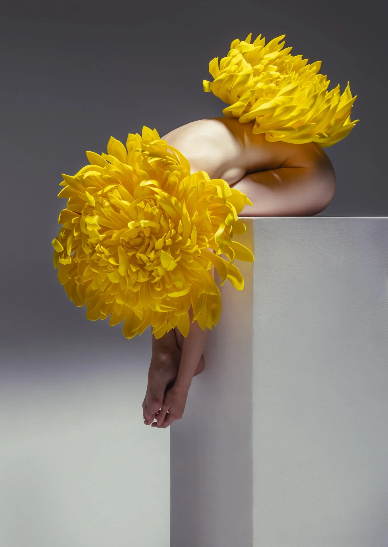 A person with large yellow flower headpieces on their head and around their waist, leaning over a white pedestal against a gray background.