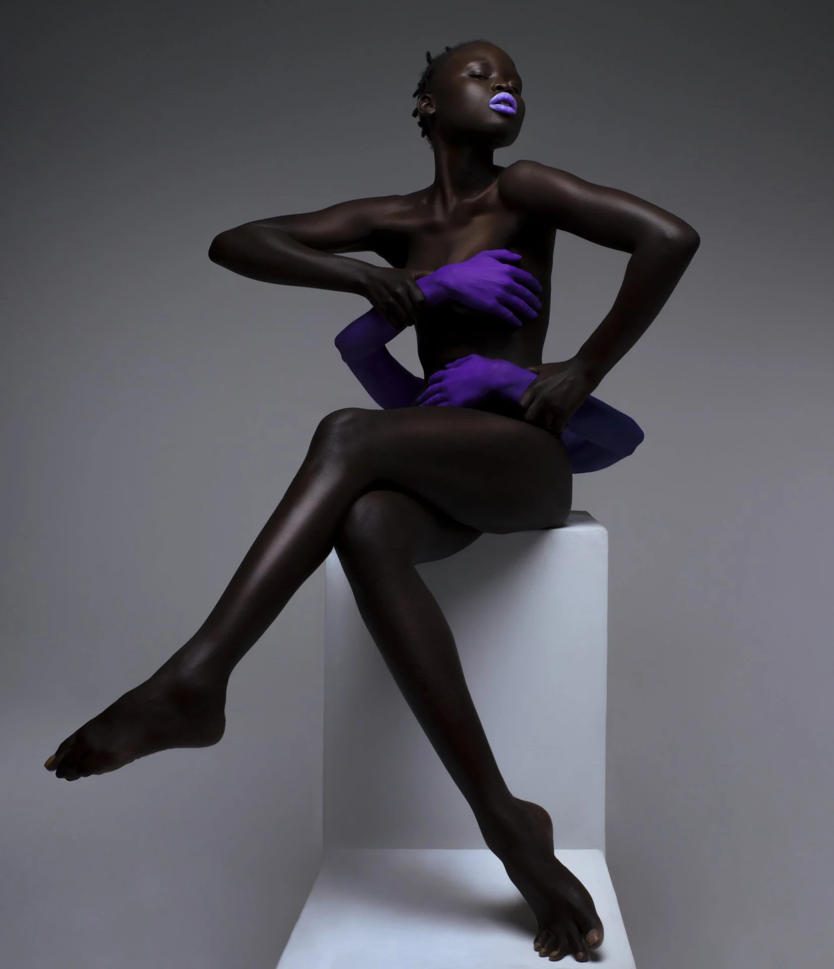 A woman with dark skin and purple lipstick sitting on a white block, with her arms crossed over her chest and legs crossed, wearing purple gloves.