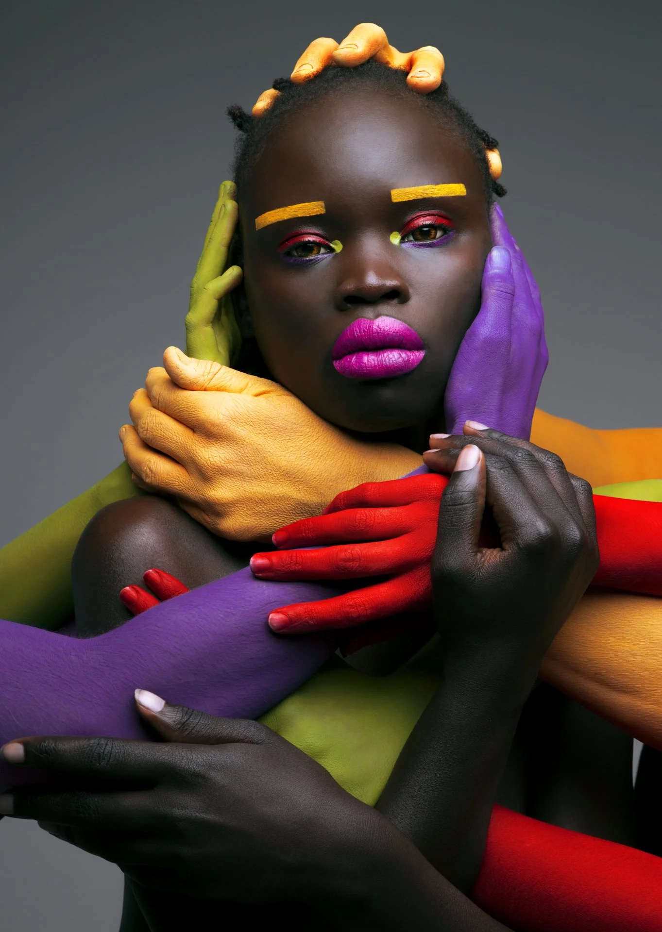 A portrait of a woman with multicolored painted hands, face, and eyebrows, emphasizing vibrant makeup and diverse skin tones, with her hands gently carved around her face.