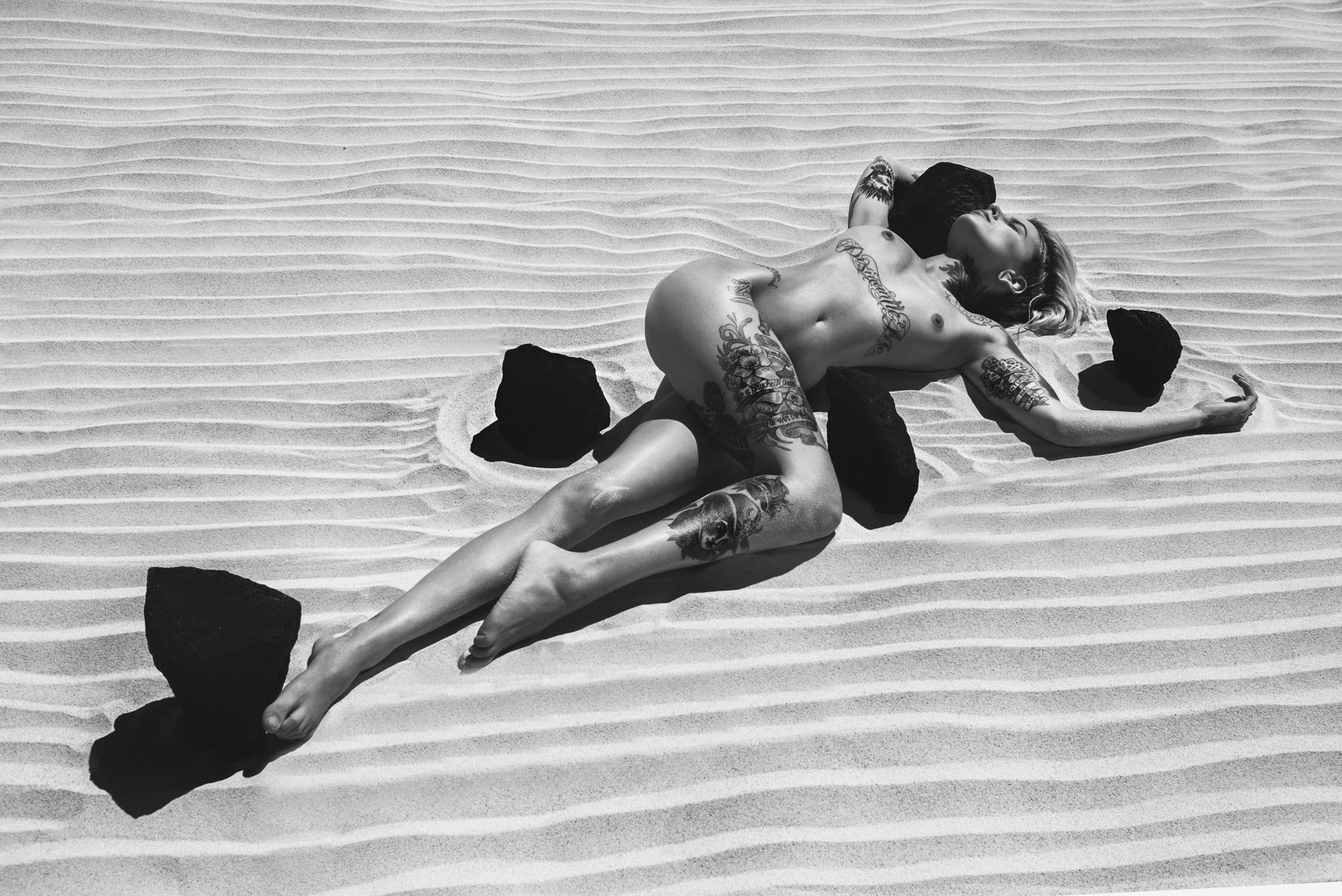 A tattooed woman lying nude on sand, surrounded by large black rocks, in a desert or beach setting.