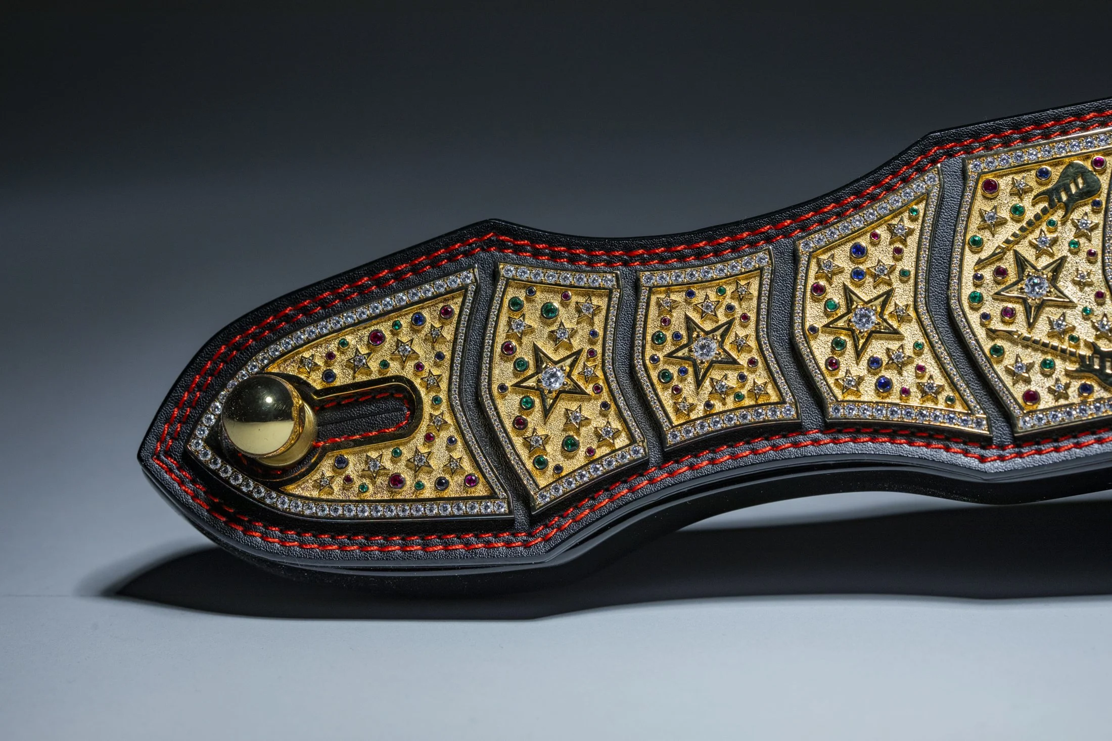 Close-up of a decorated championship belt with gold, diamonds, and colored gemstones, featuring detailed star patterns and red stitching along the black leather strap.