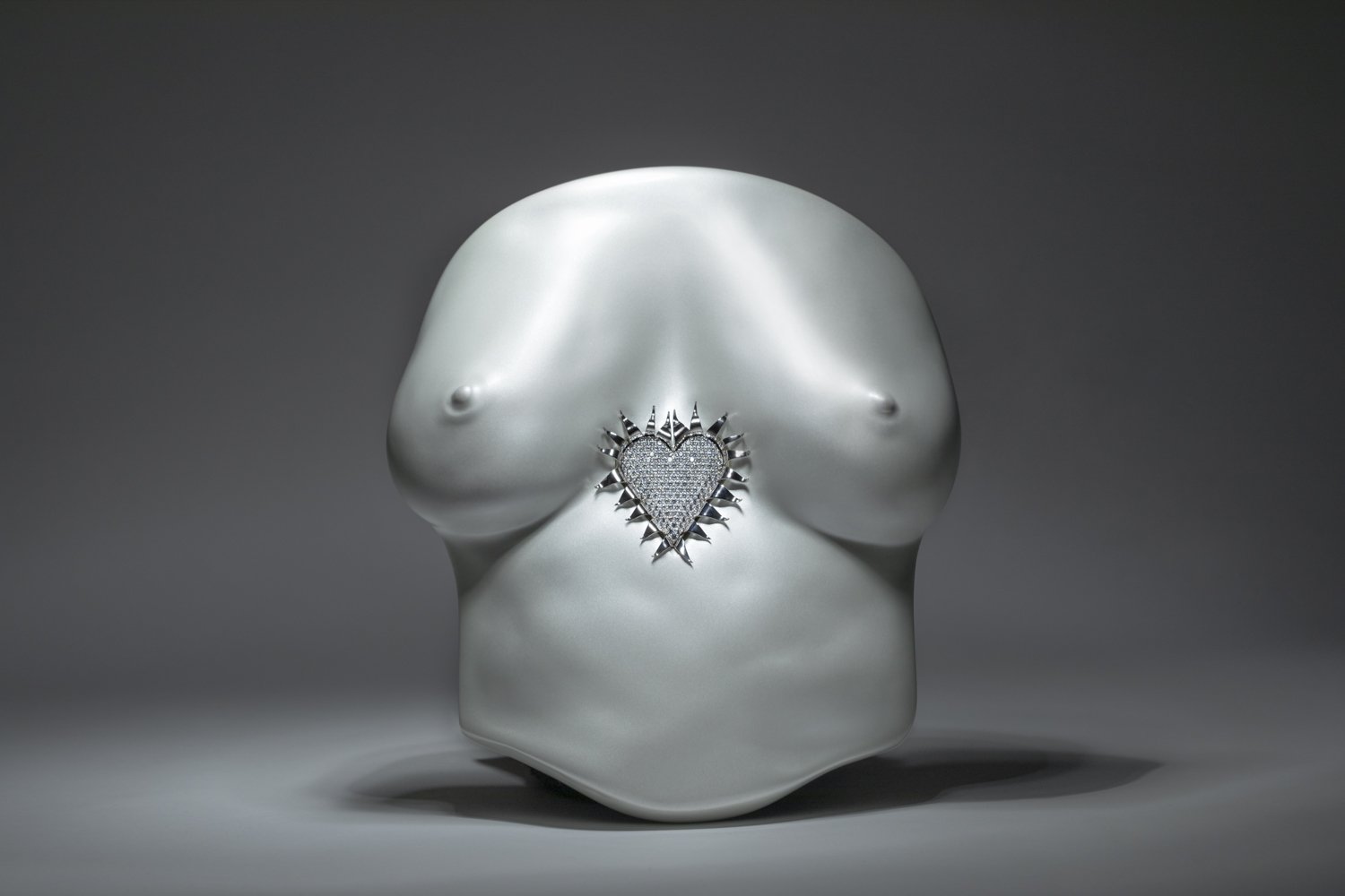 A silver, heart-shaped pendant with a spiky border and embedded diamonds, placed on a smooth, abstract silver sculpture resembling a humanoid torso.