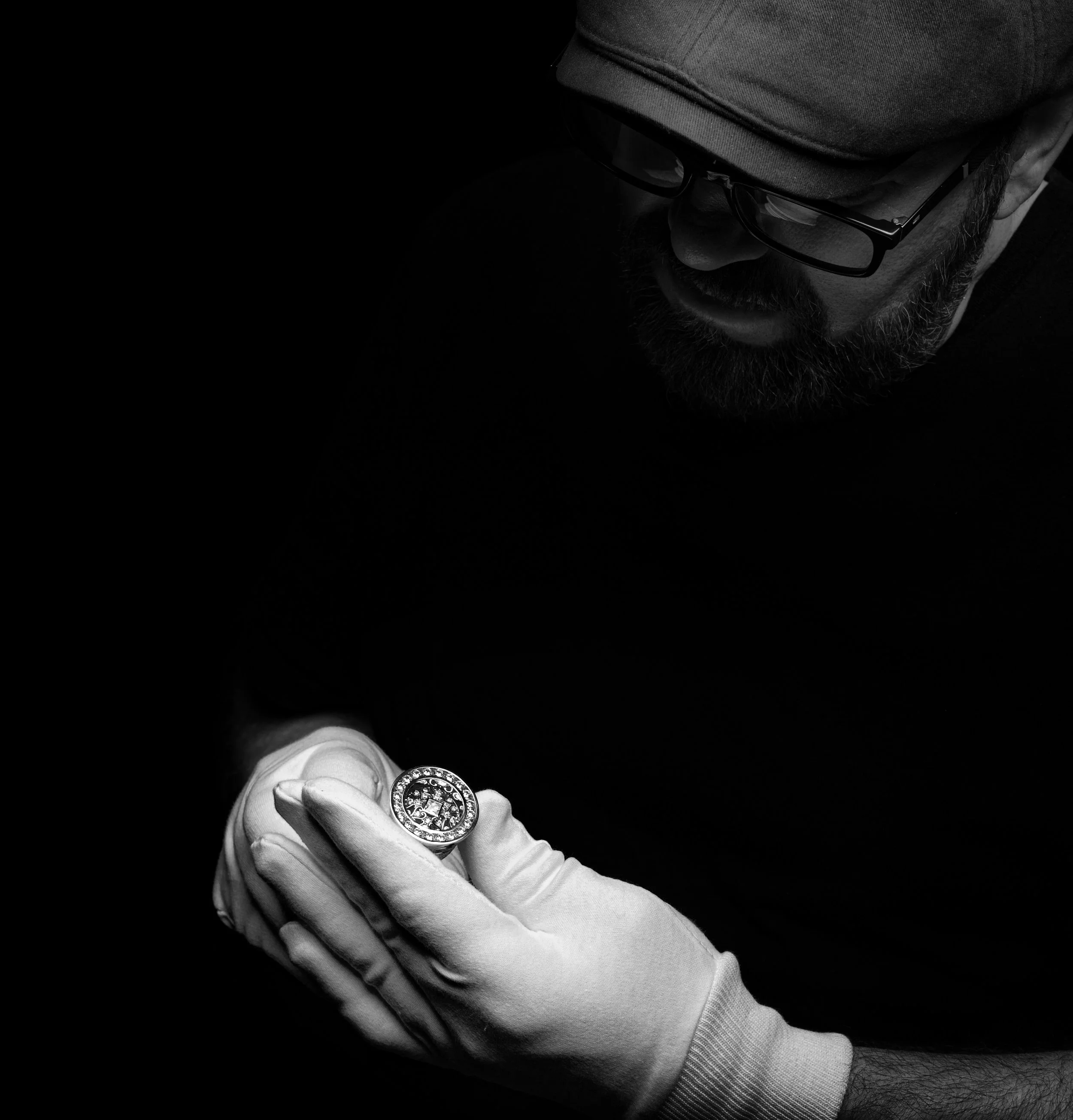 A man with glasses and a hat looking down at a large ornate ring on his finger, wearing a light-colored glove, against a dark background.