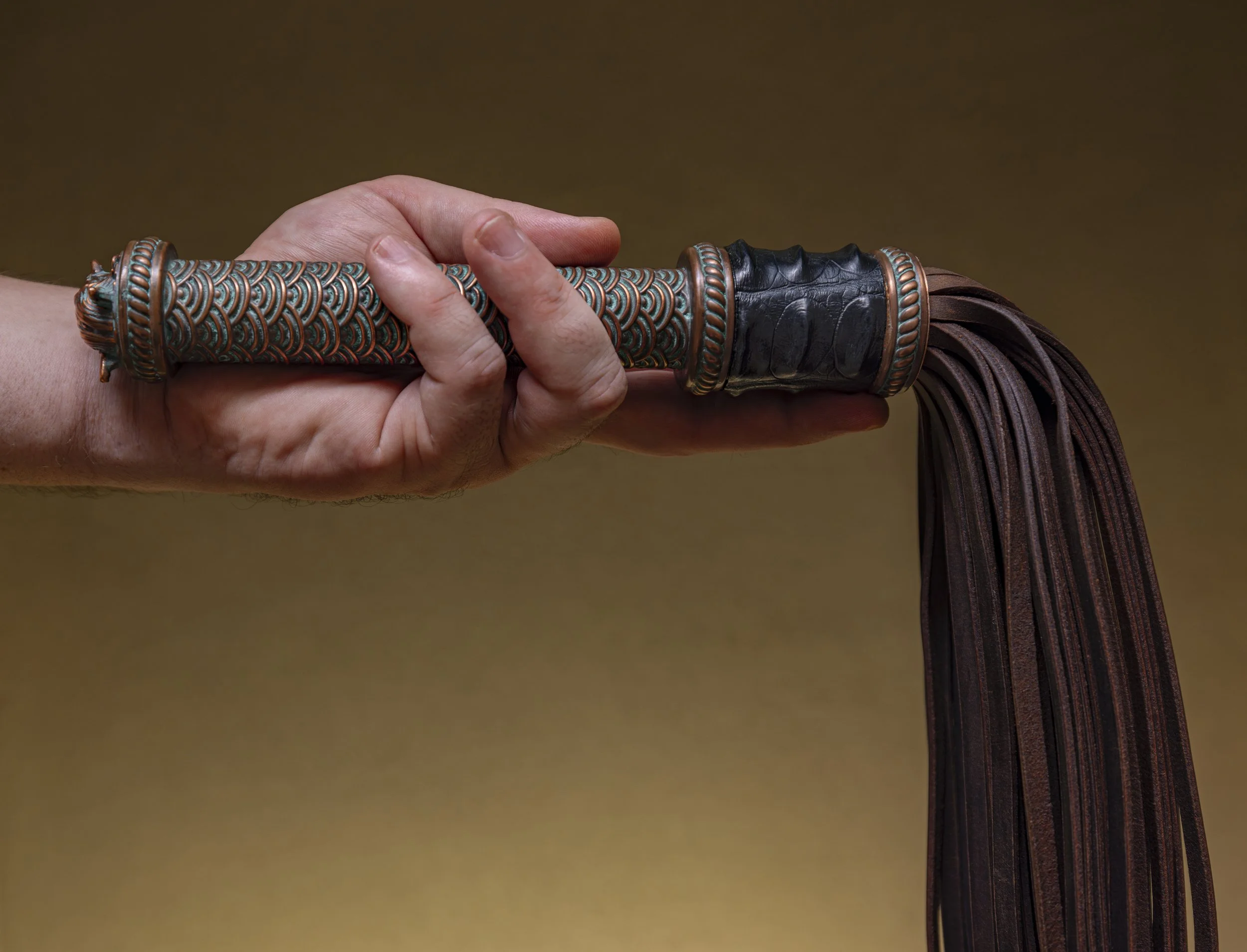 Hand holding a woven and leather decorated flogger toy with leather straps hanging down.