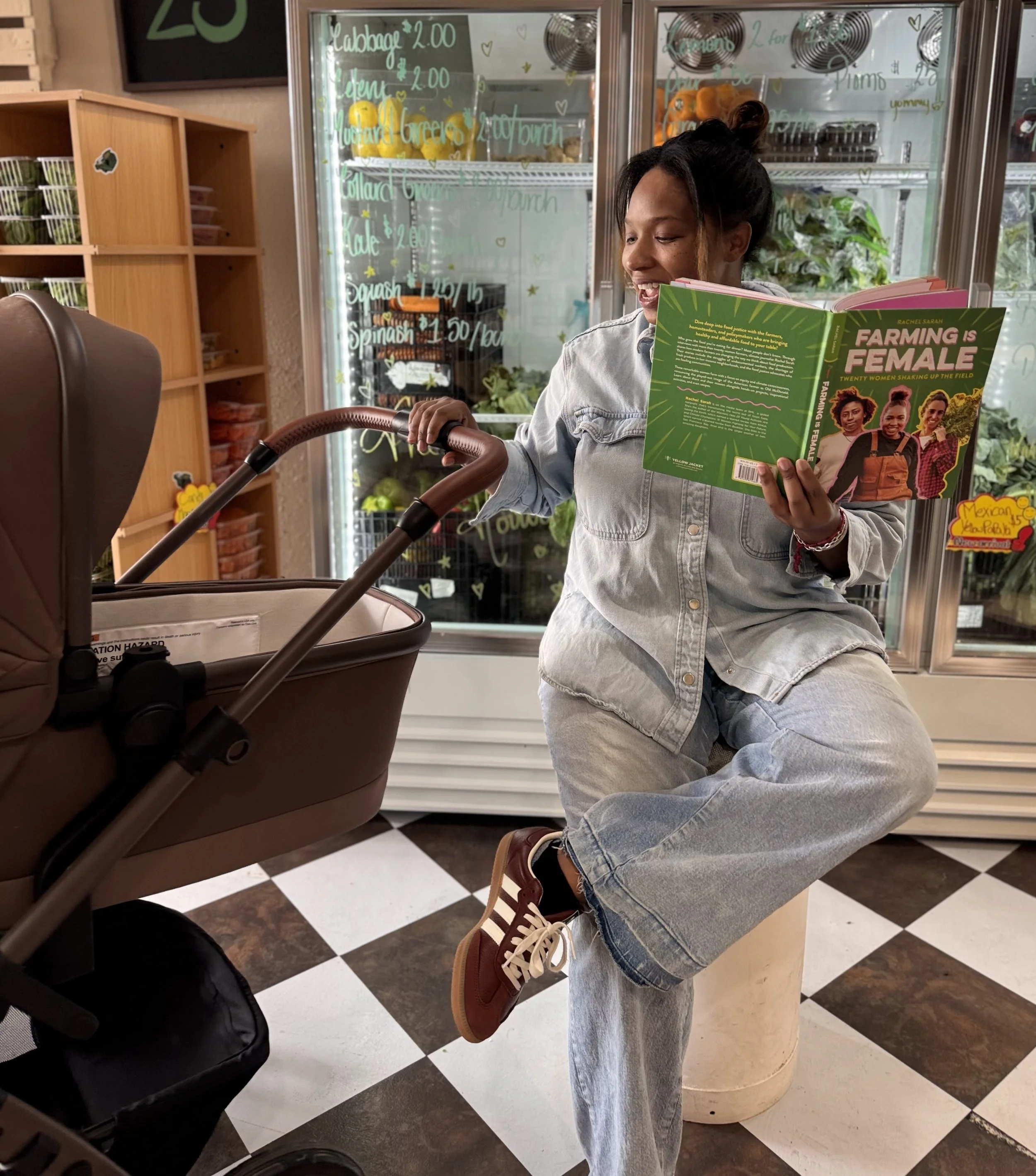A woman sits on a stool inside a grocery store, reading a book titled 'Farming is Female' while holding a baby stroller nearby. She is wearing a light denim jacket, matching jeans, and Adidas sneakers.