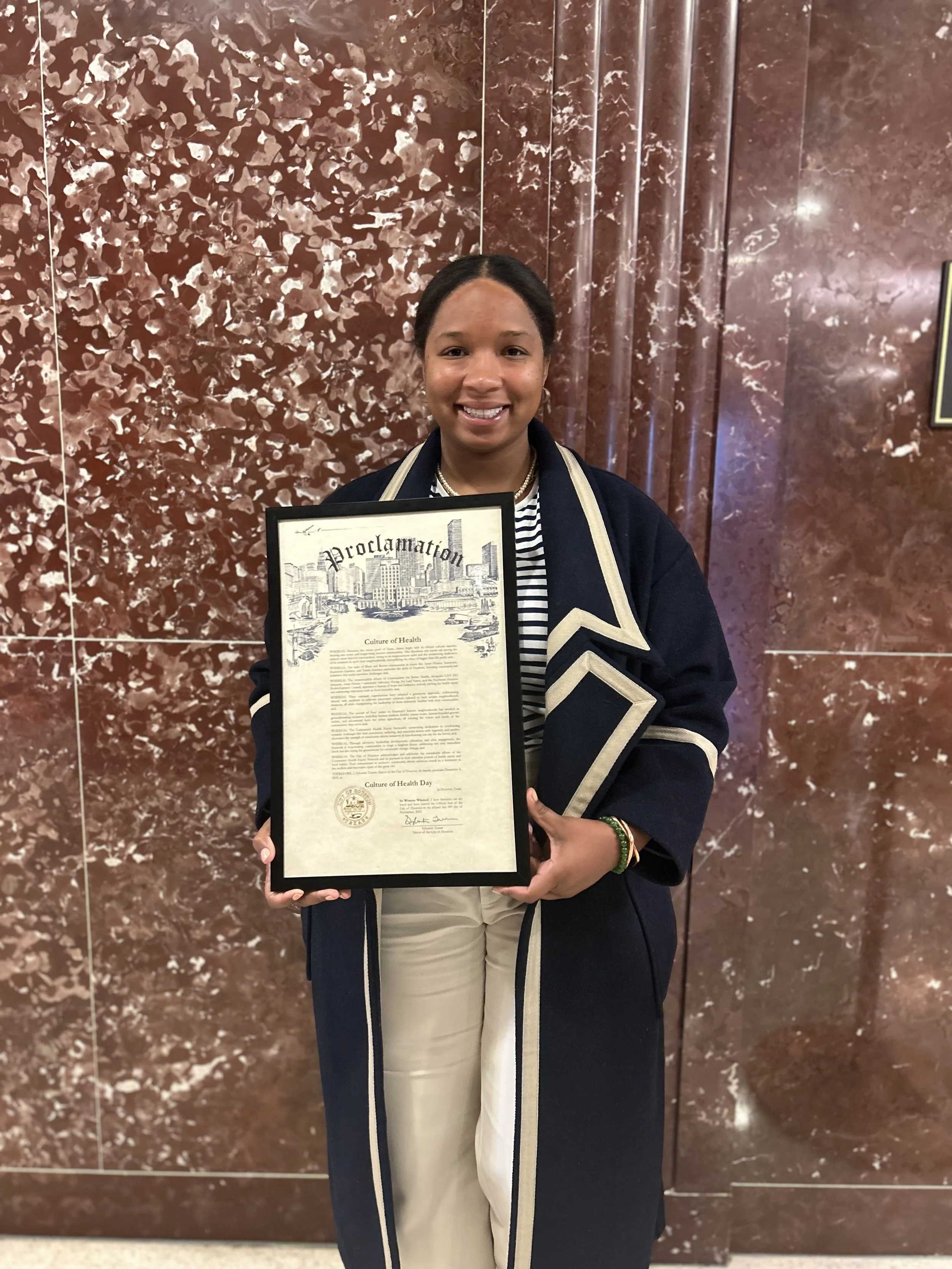 A young woman smiling while holding a framed document titled 'Proclamation' in front of a brown marble wall.