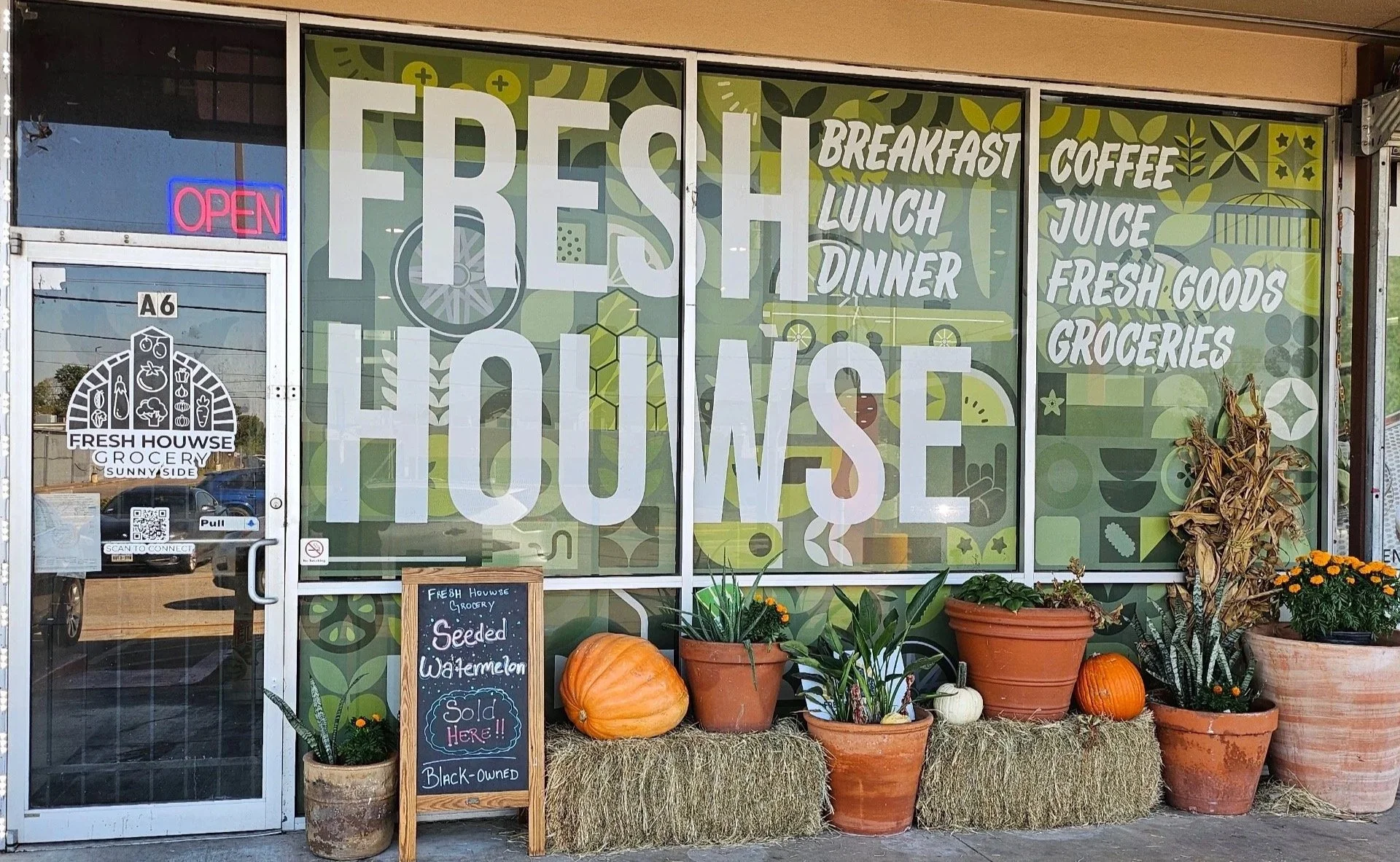 Grocery store storefront with large windows featuring 'Fresh House' signage, decorated with pumpkins, potted plants, and hay bales outside, with a neon 'Open' sign and a chalkboard advertising seedless watermelons.