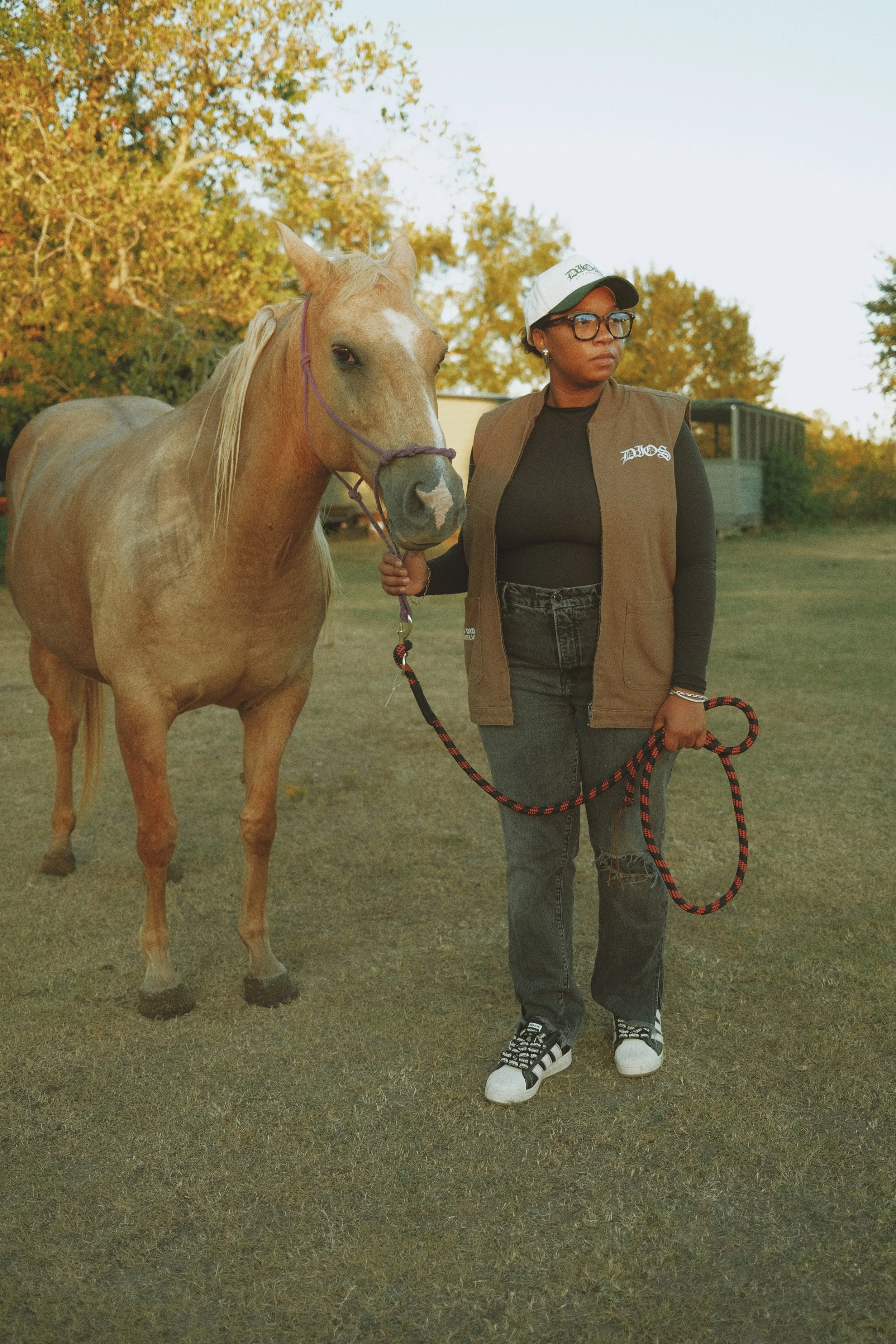 A woman standing outdoors holding a leash attached to a light brown horse, with trees in the background during daytime.