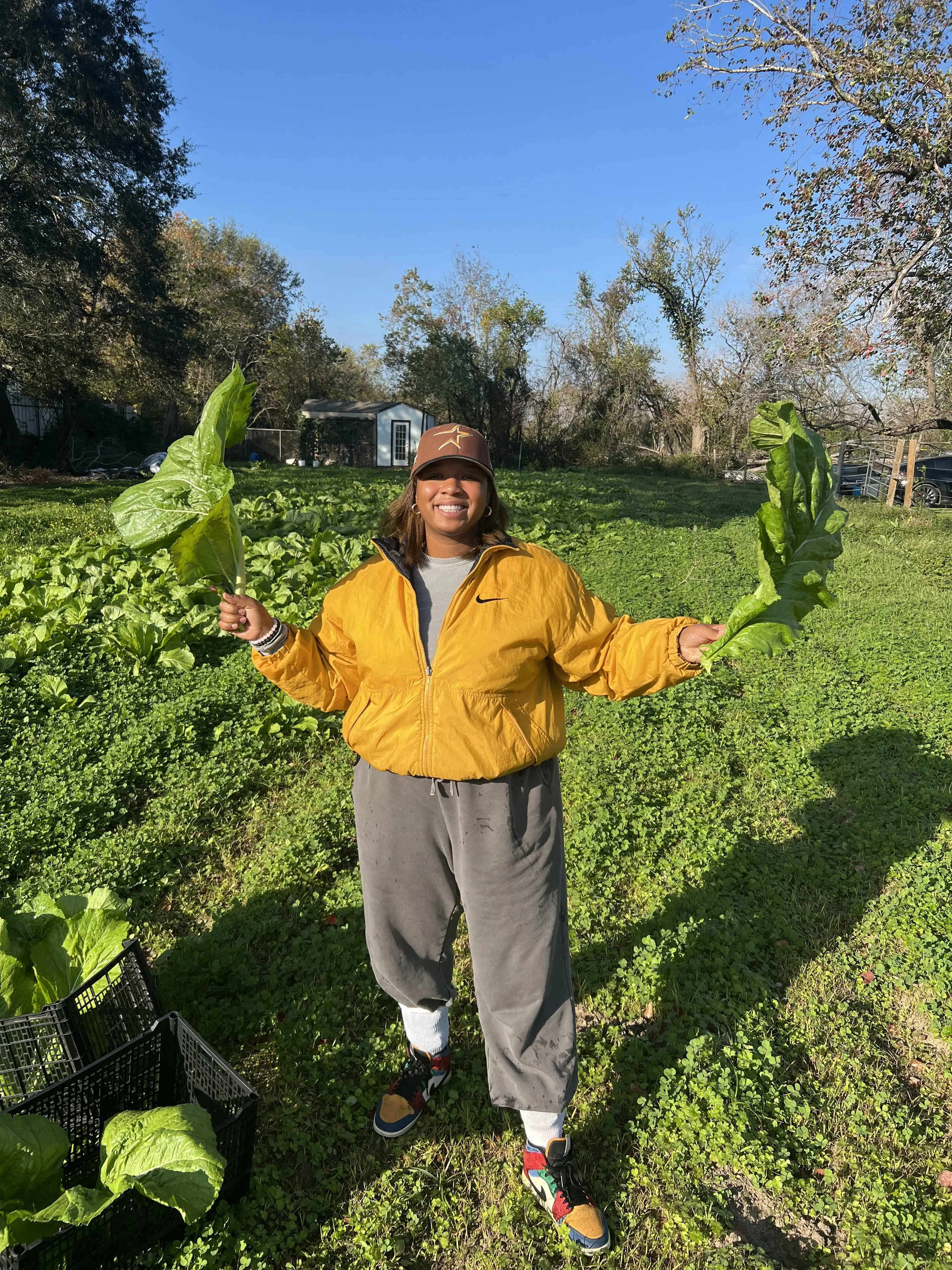 Smiling woman standing in a garden, holding large leafy greens in each hand, wearing a yellow jacket, gray pants, colorful sneakers, and a cap.