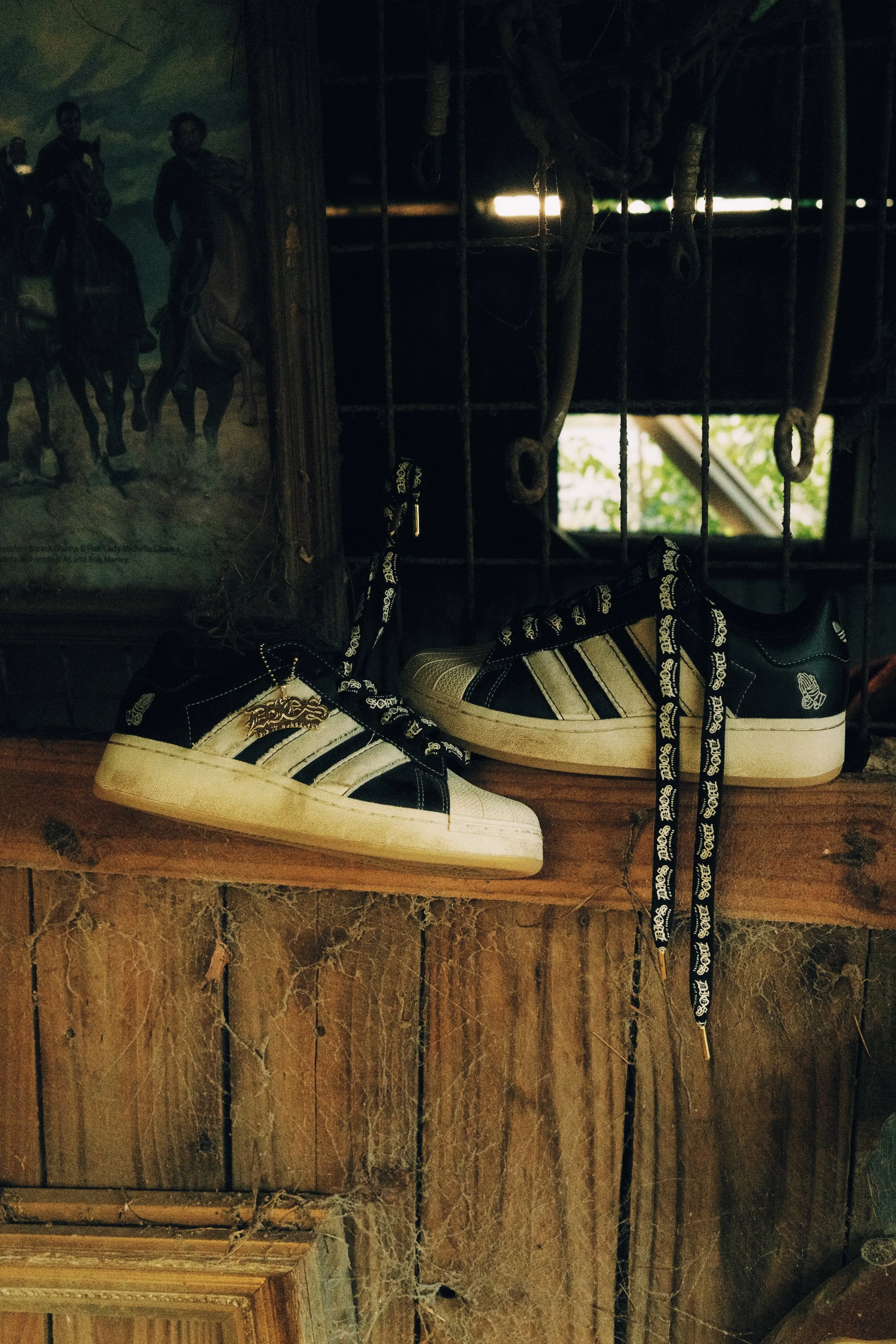 A pair of black and white Adidas sneakers on a wooden shelf, surrounded by cobwebs, with a painting and a window in the background.