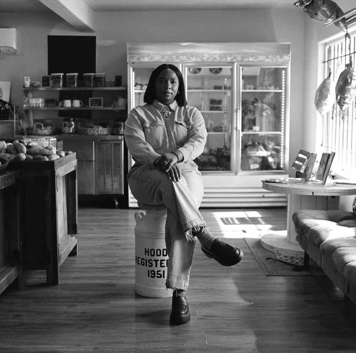 A woman sitting on a barrel in a cozy store, surrounded by shelves of goods and a window with sunlight.