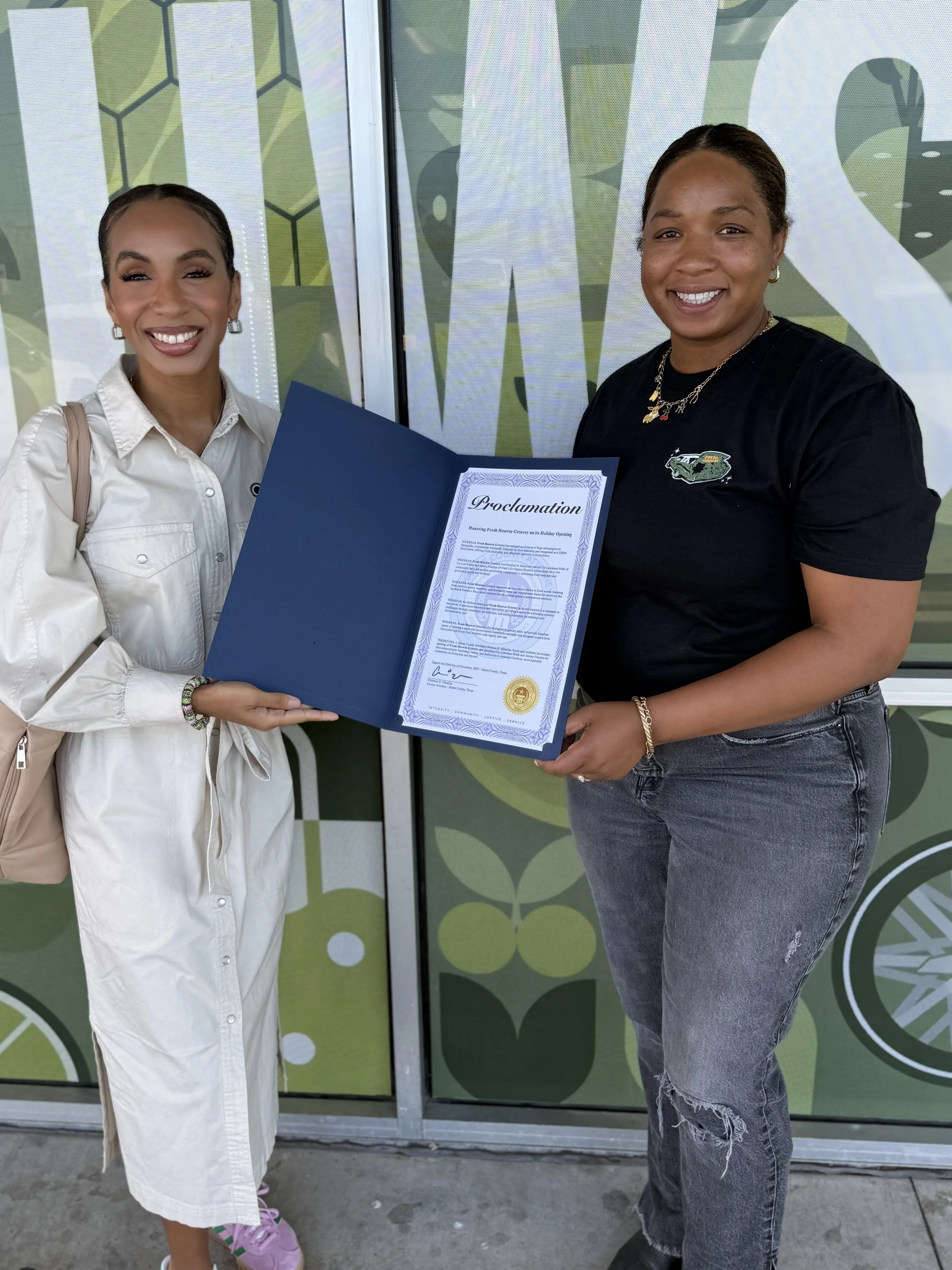 Two women smiling, holding a large proclamation certificate, standing in front of a green patterned backdrop.