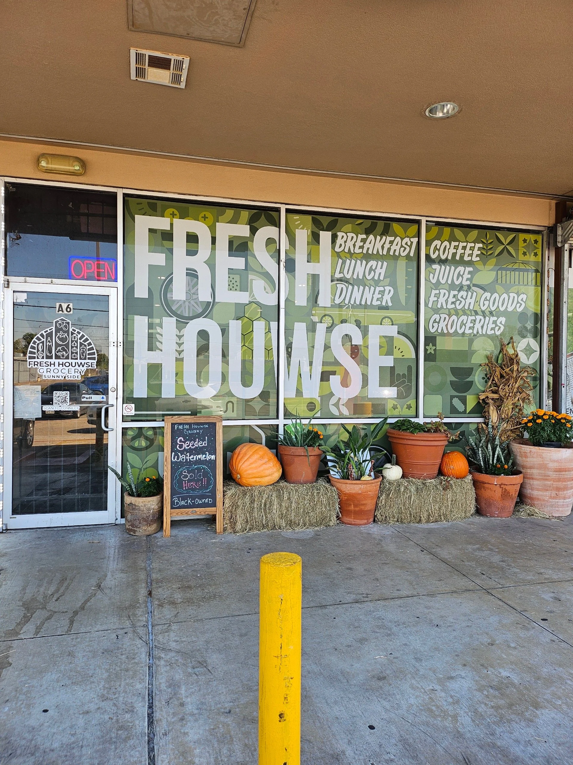 Exterior of a grocery store called Fresh House with large window decals advertising breakfast, lunch, dinner, coffee, juice, fresh goods, and groceries. The storefront is decorated with pumpkins, potted plants, hay bales, and a chalkboard sign indicating the sale of seeded watermelon, owned by Black.