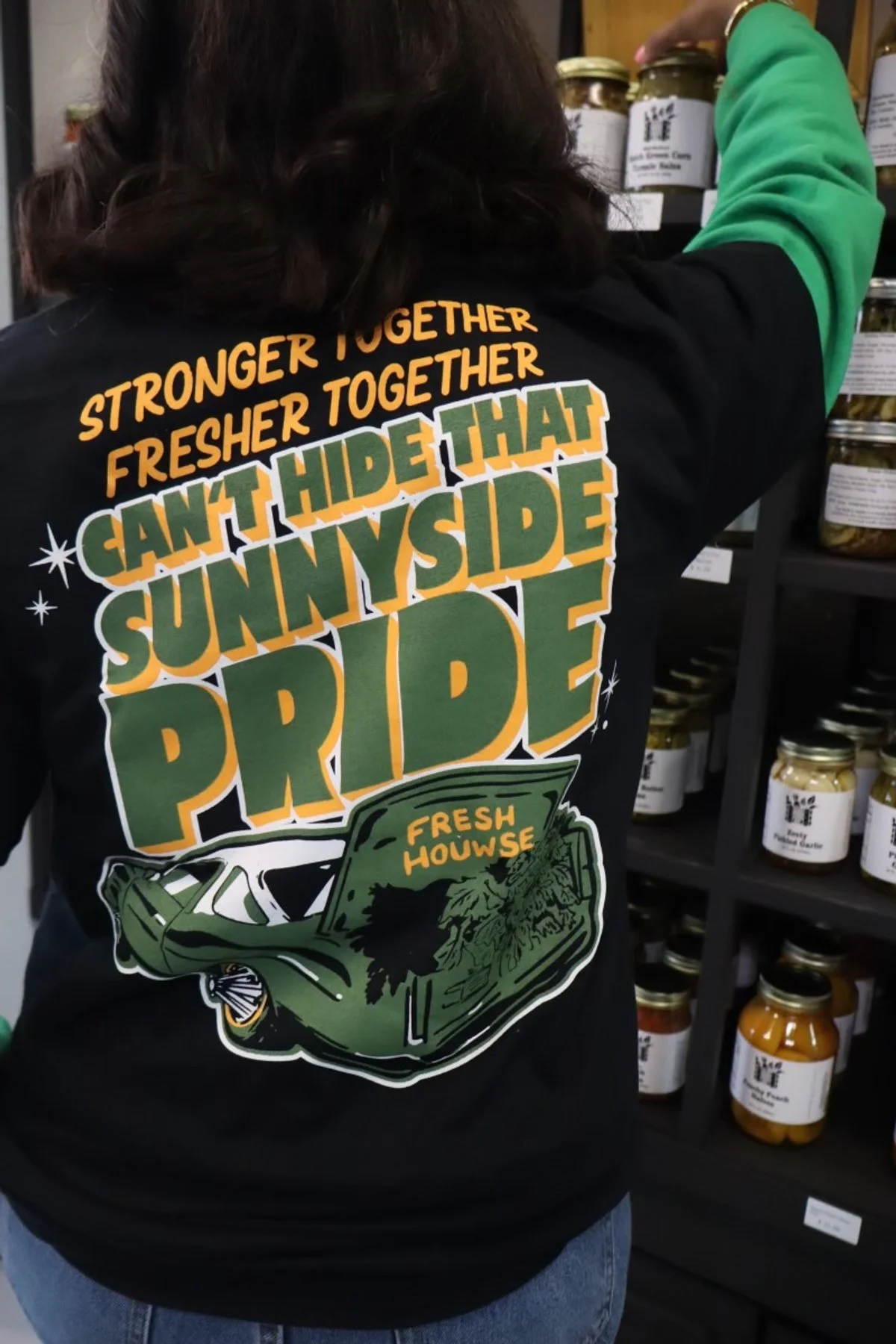 Person wearing a black T-shirt with colorful text and graphics, reaching for jar on shelf. The shirt reads, 'Stronger together, fresher together, can't hide that sunnyside pride' and features a graphic of a car and a grocery produce bag with the words 'Fresh House'.