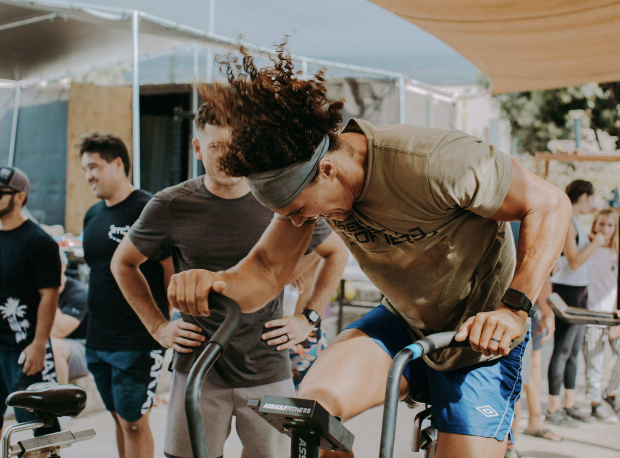 Man wearing headband and watch, bent over, pushing pedals on a stationary exercise bike during a workout at an outdoor fitness event, with other people gathered around him.