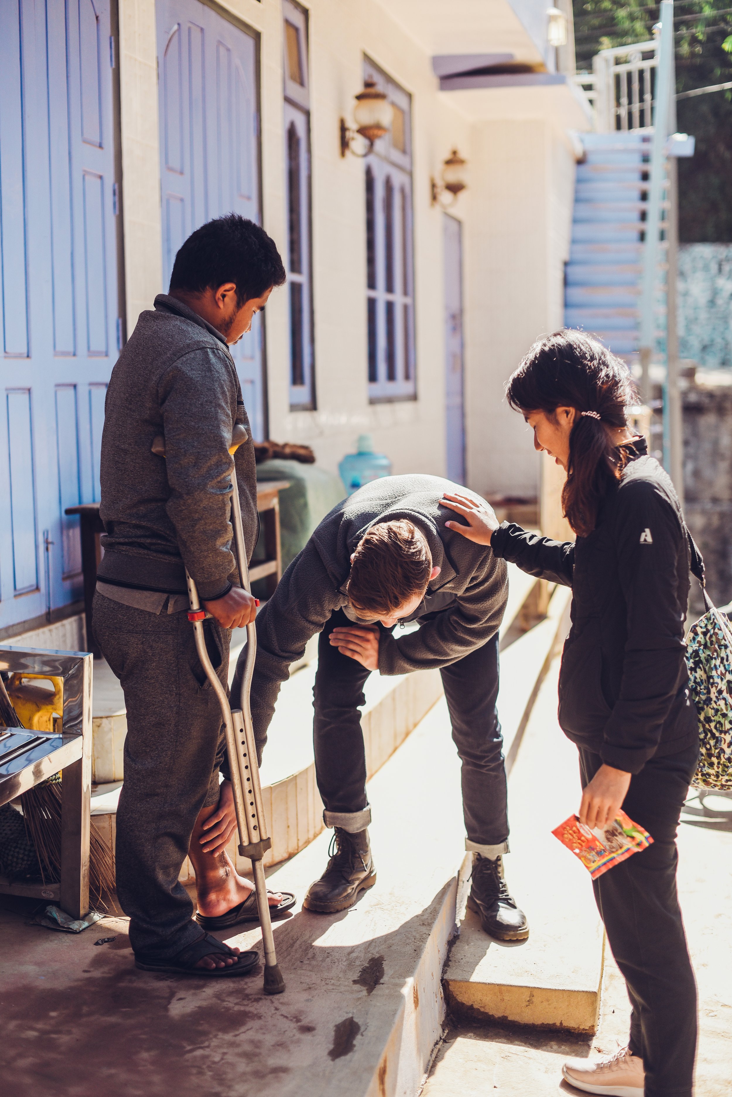 A person with a crutch leaning over and other two people comforting him outside near a building.
