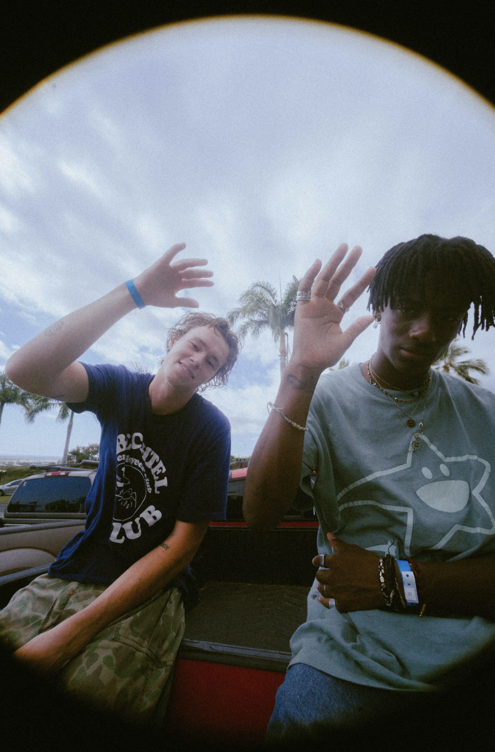 Two young men waving, standing outdoors with palm trees and a partly cloudy sky in the background.