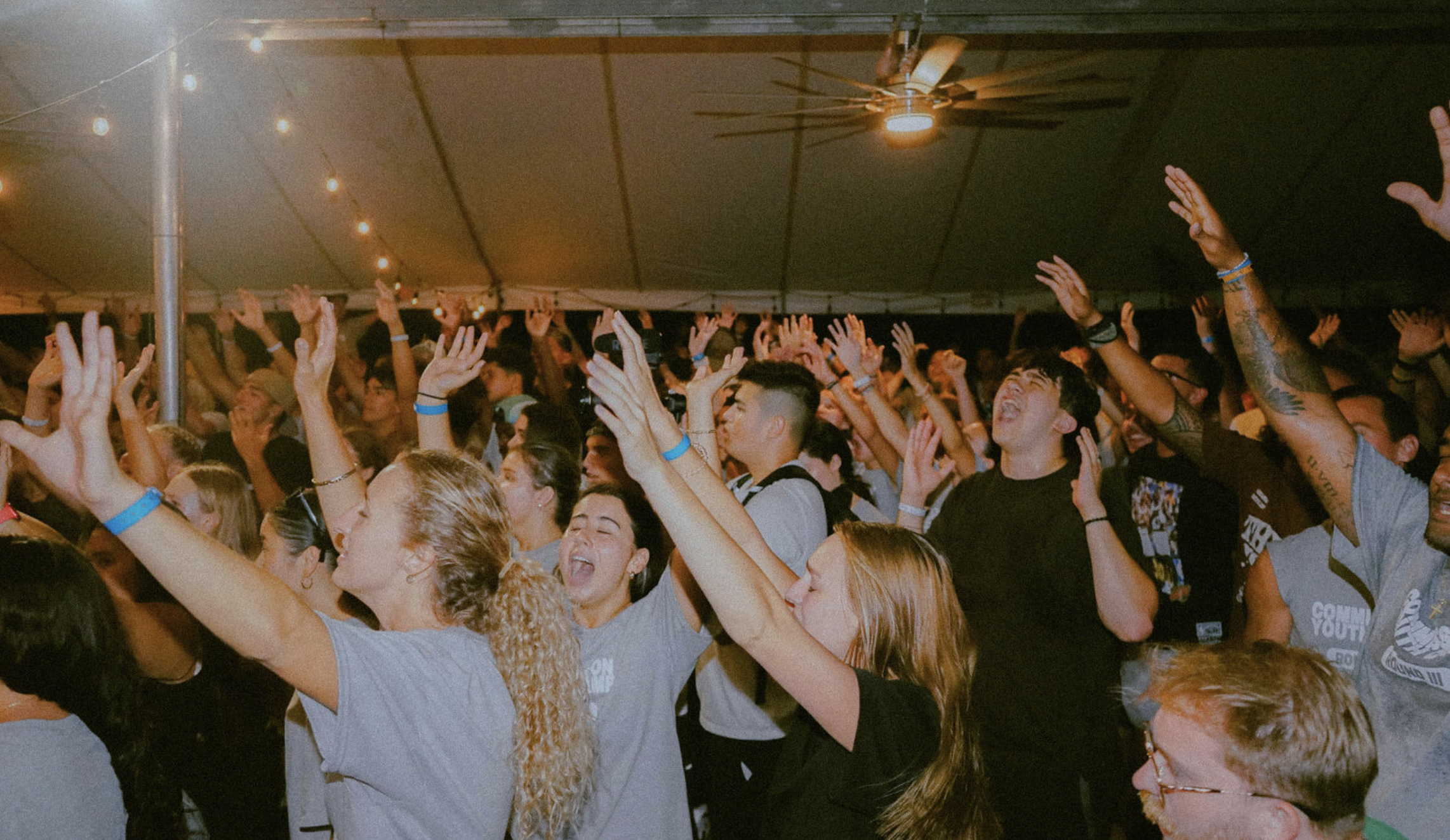 Crowd of people at a concert or event inside a tent, raising their arms and enjoying music with string lights and a ceiling fan overhead.