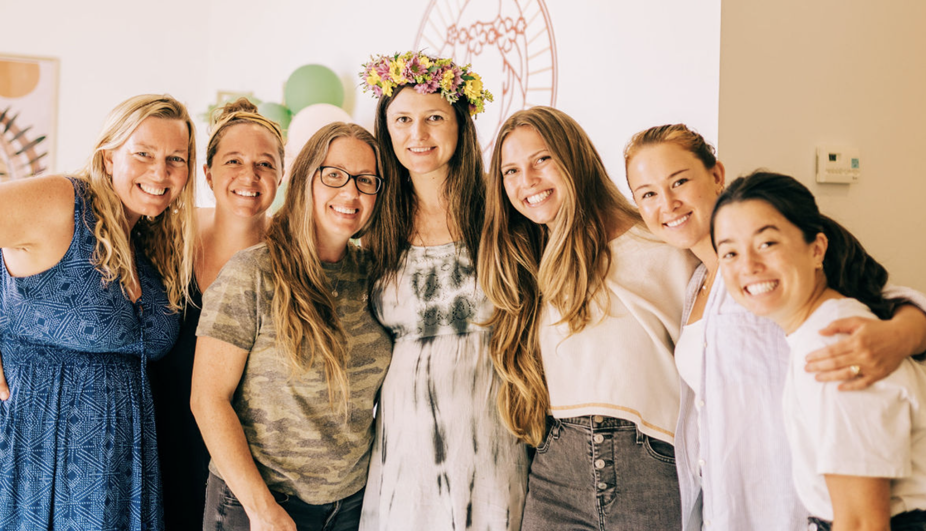 Group of women celebrating at a party, one wearing a flower crown, decorated with balloons and wall art in the background.