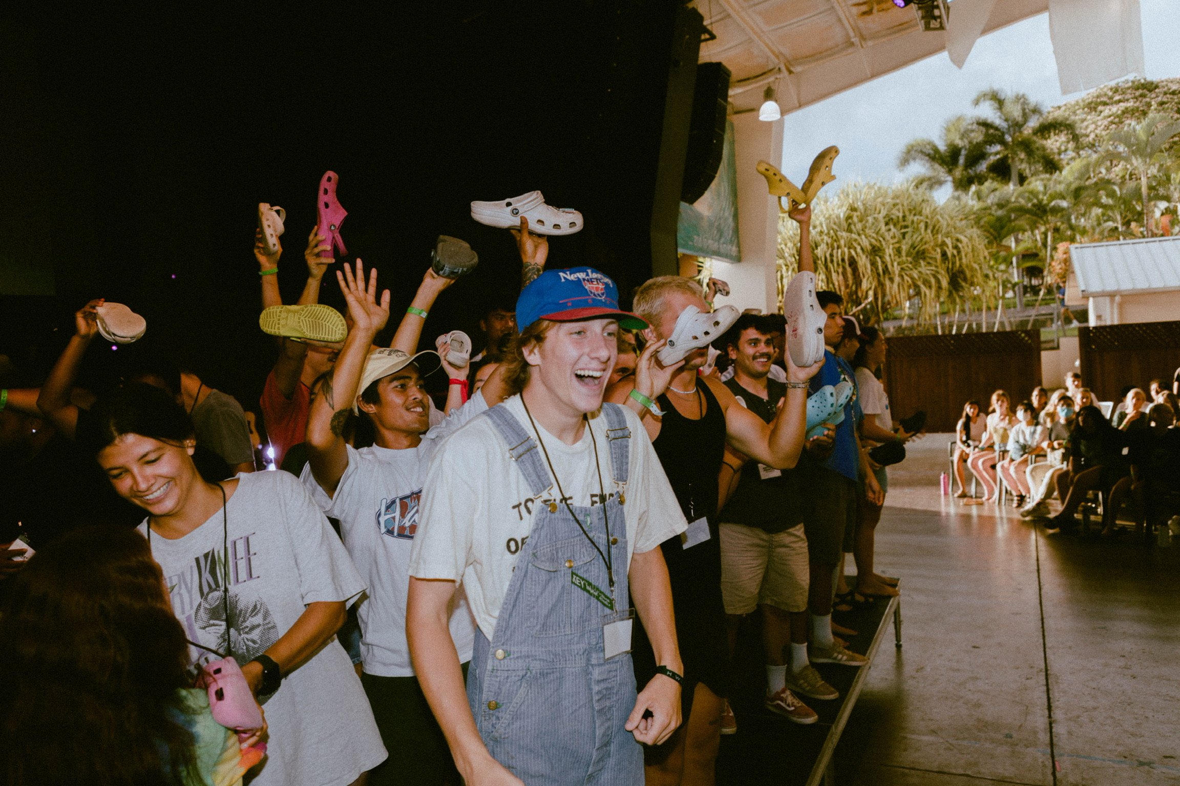 Group of people holding colorful shoes above their heads at an indoor event with a stage and outdoor scenery in the background.
