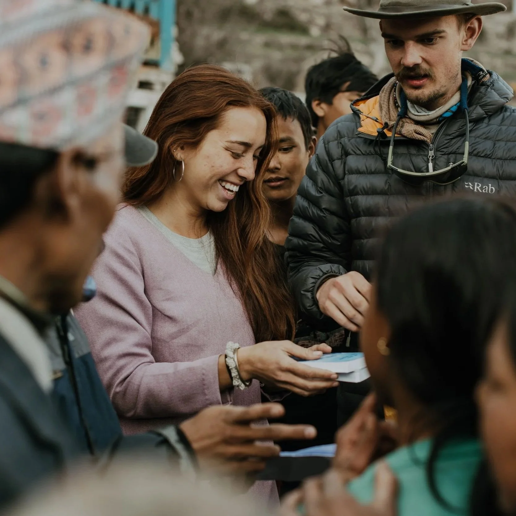 Group of diverse people gathered outdoors, smiling and looking at a small booklet or pamphlet.