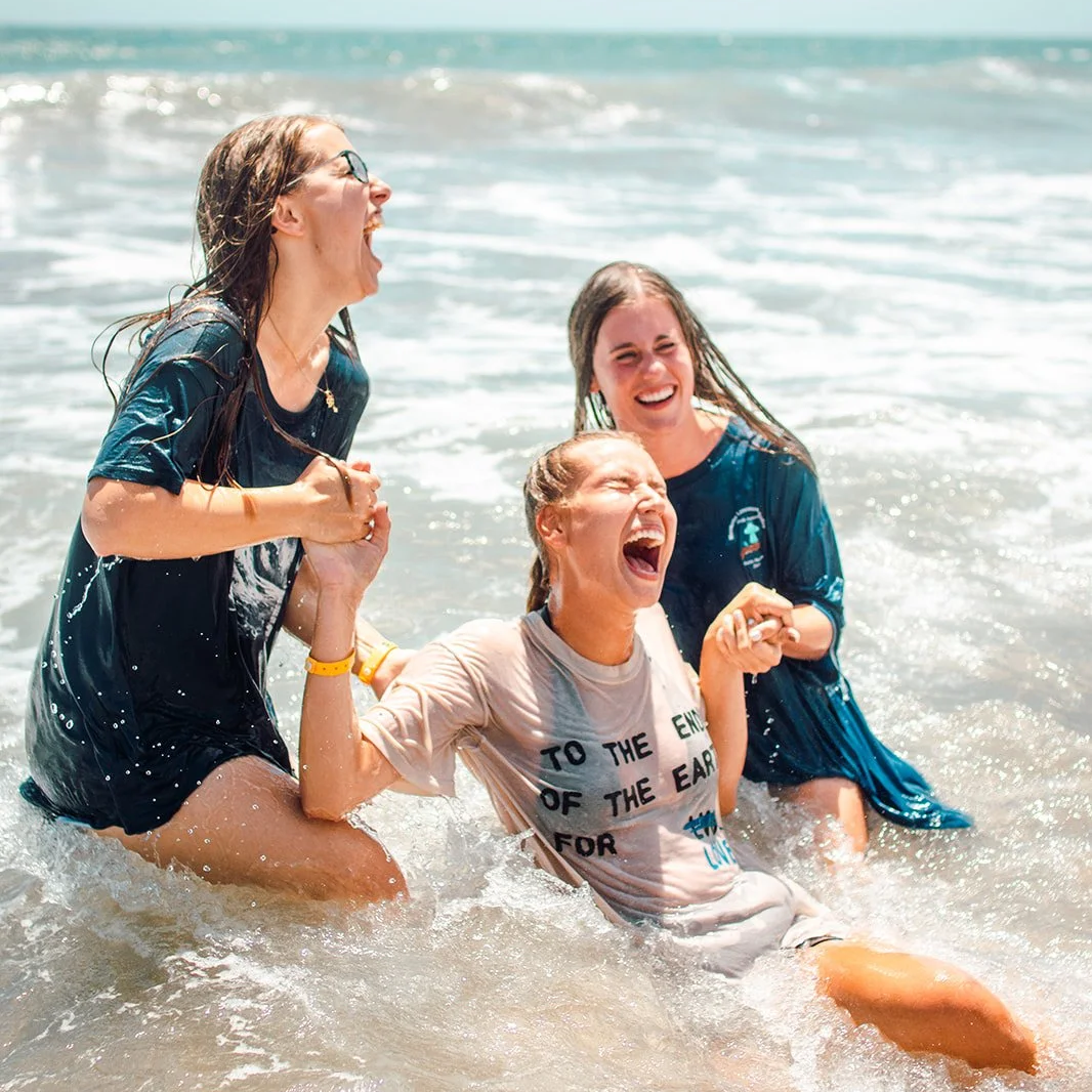 Three women laughing and playing in the ocean water, holding hands in a joyful moment.