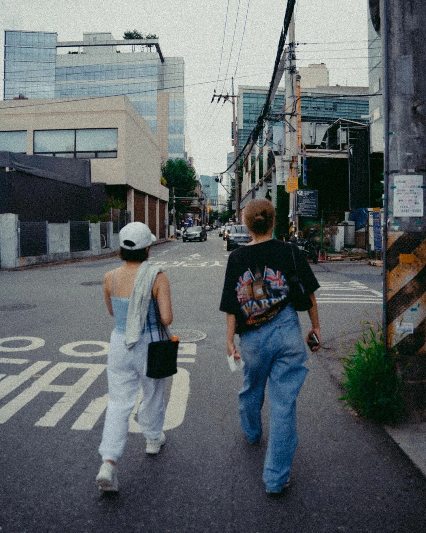 Two women walking on an urban street with tall buildings and power lines overhead.
