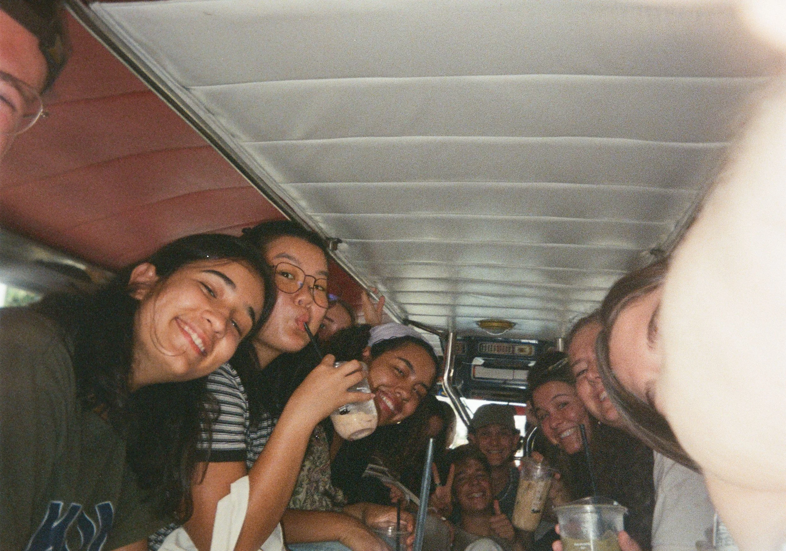 A group of young women and one young man sitting closely together inside a bus, smiling and enjoying drinks with straws, with some making playful faces and looking at the camera.