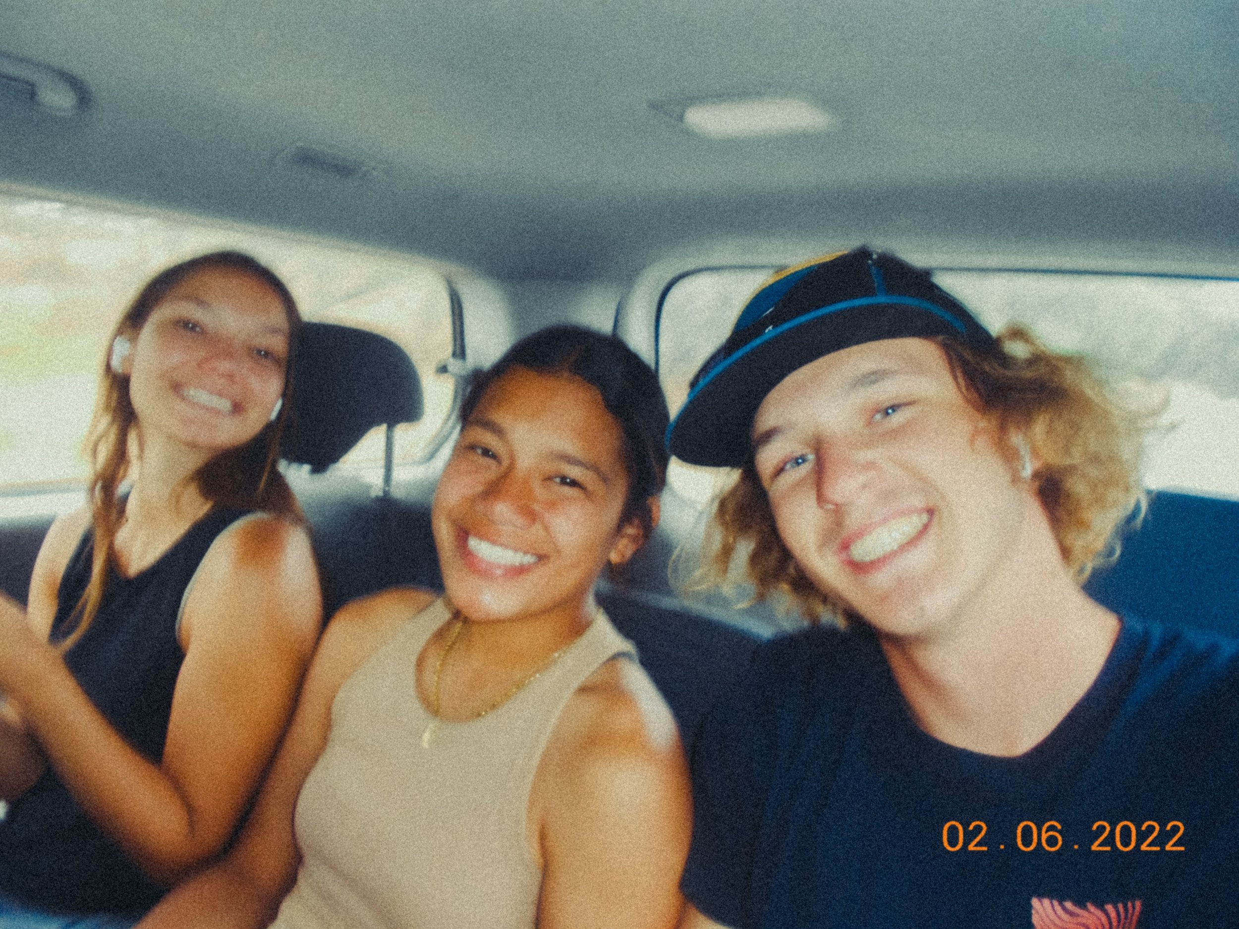 Three young friends smiling and posing for a selfie in a car, with a date stamp of February 6, 2022 in the corner.