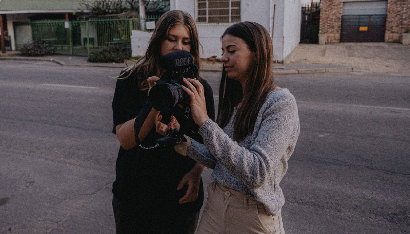 Two women standing on a street, looking at a camera. One woman holds a professional camera with a microphone attached, and the other woman points at the camera's screen.