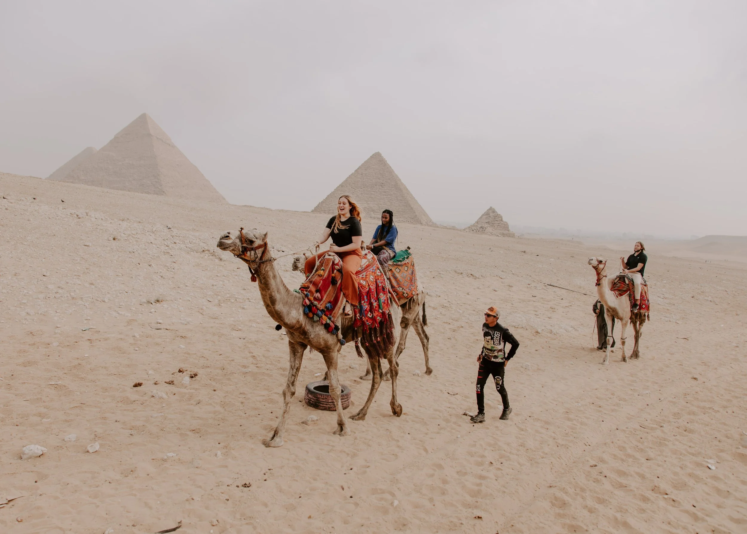 People riding camels in the desert with pyramids in the background.