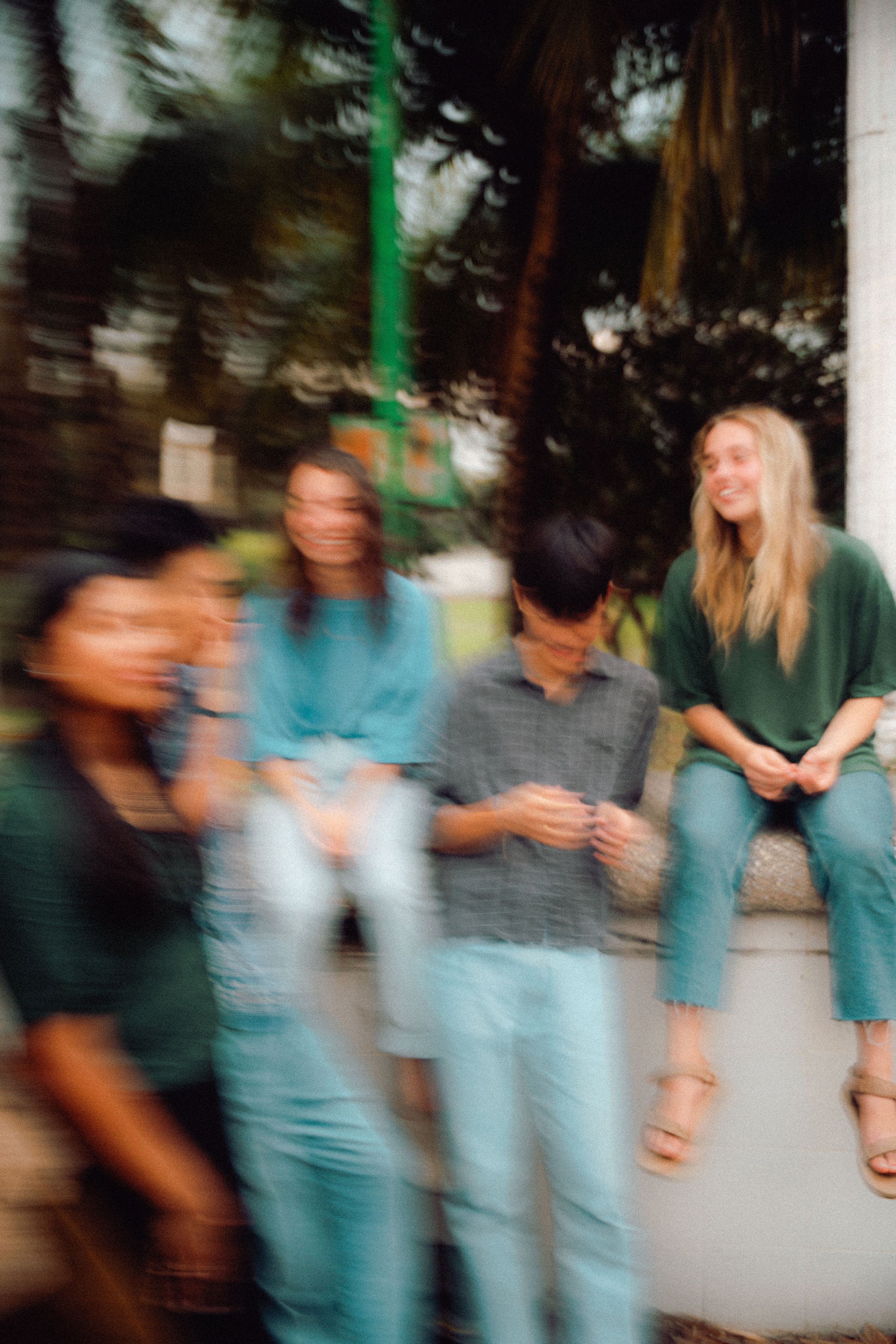Blurry photo of five young people talking and laughing outdoors during daytime, with trees and a green pole in the background.