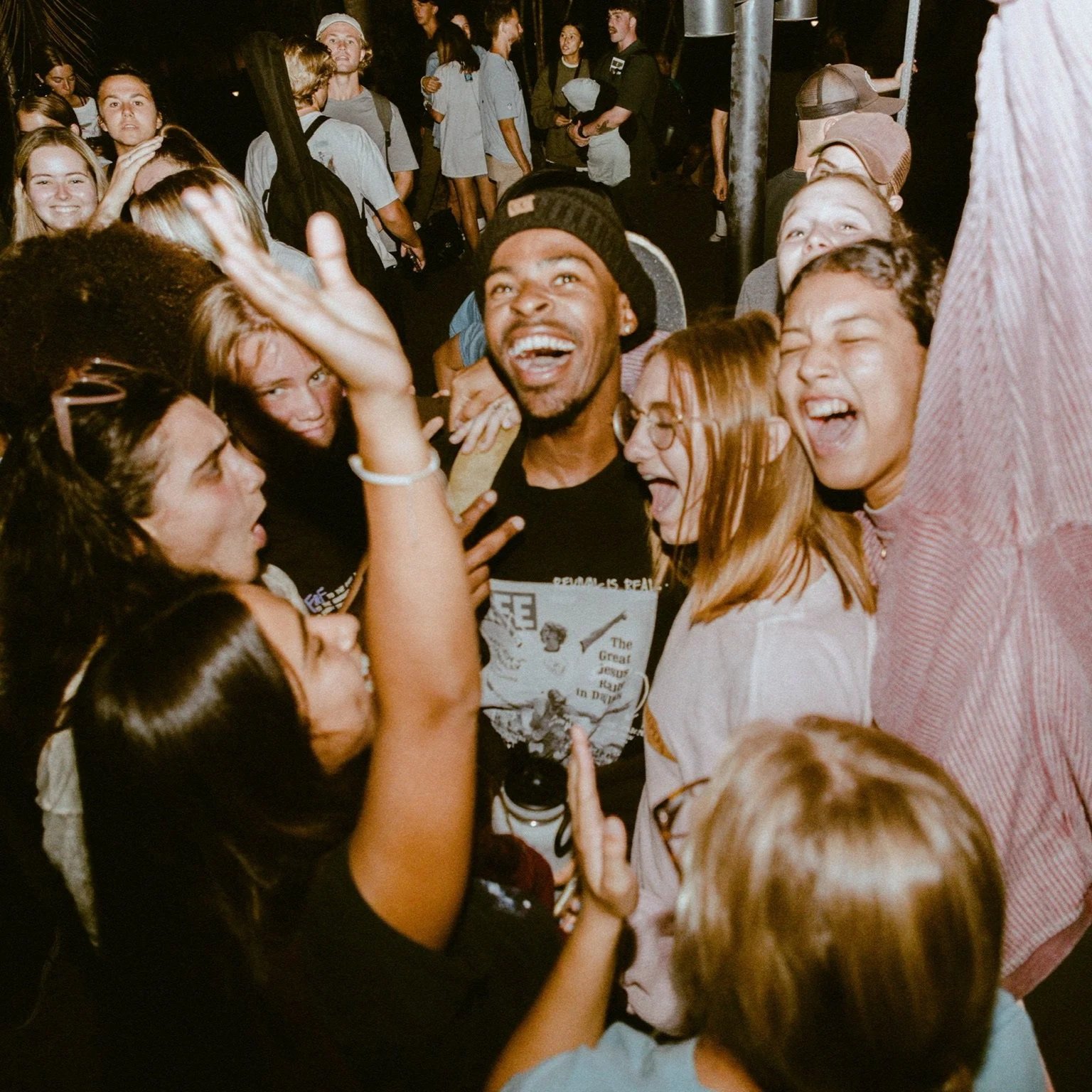 A diverse group of young people, smiling and laughing, gathered closely in a lively crowd at night.