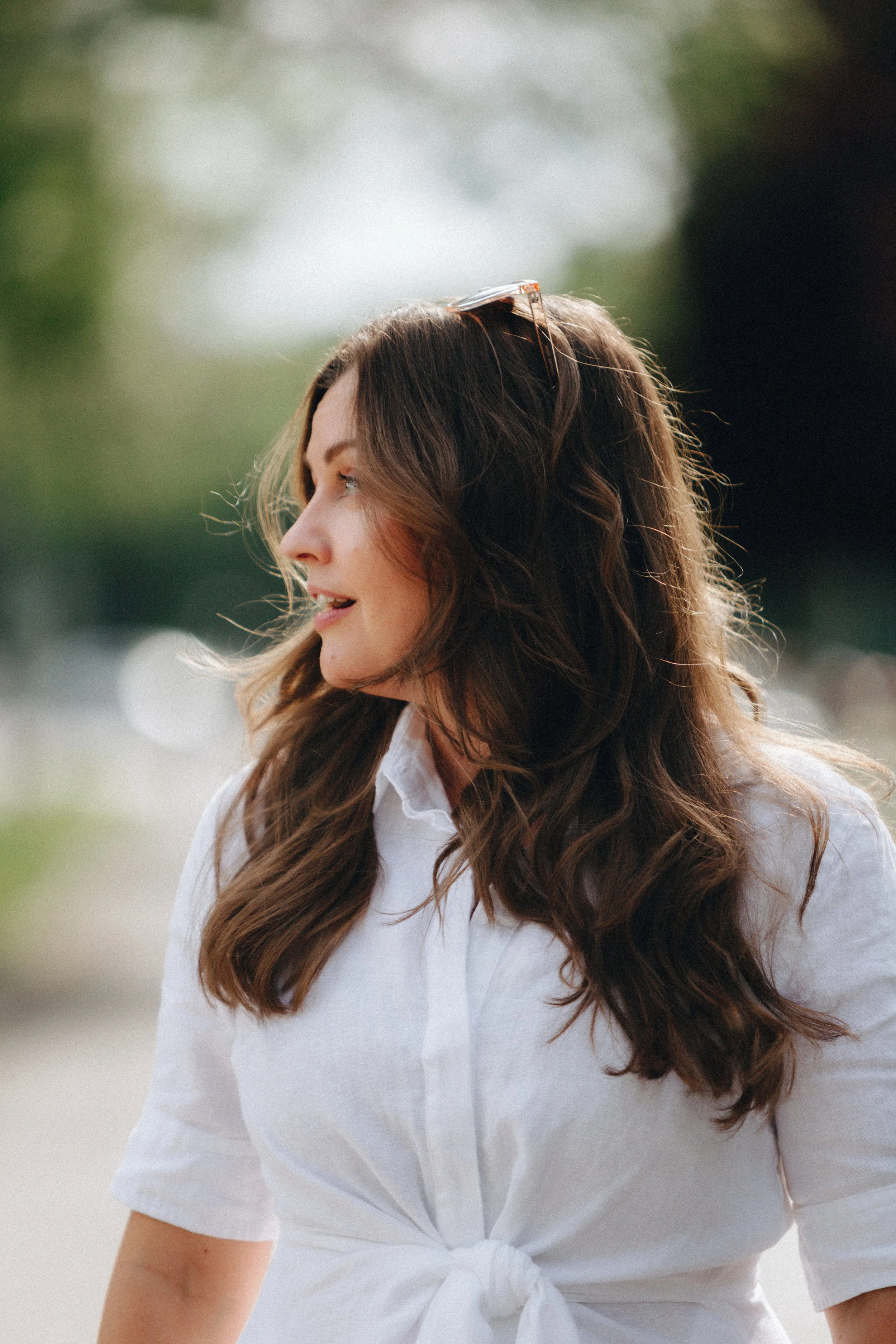 Side profile of a woman with long, wavy brown hair, wearing sunglasses on her head and a white front-tied shirt, outdoors.