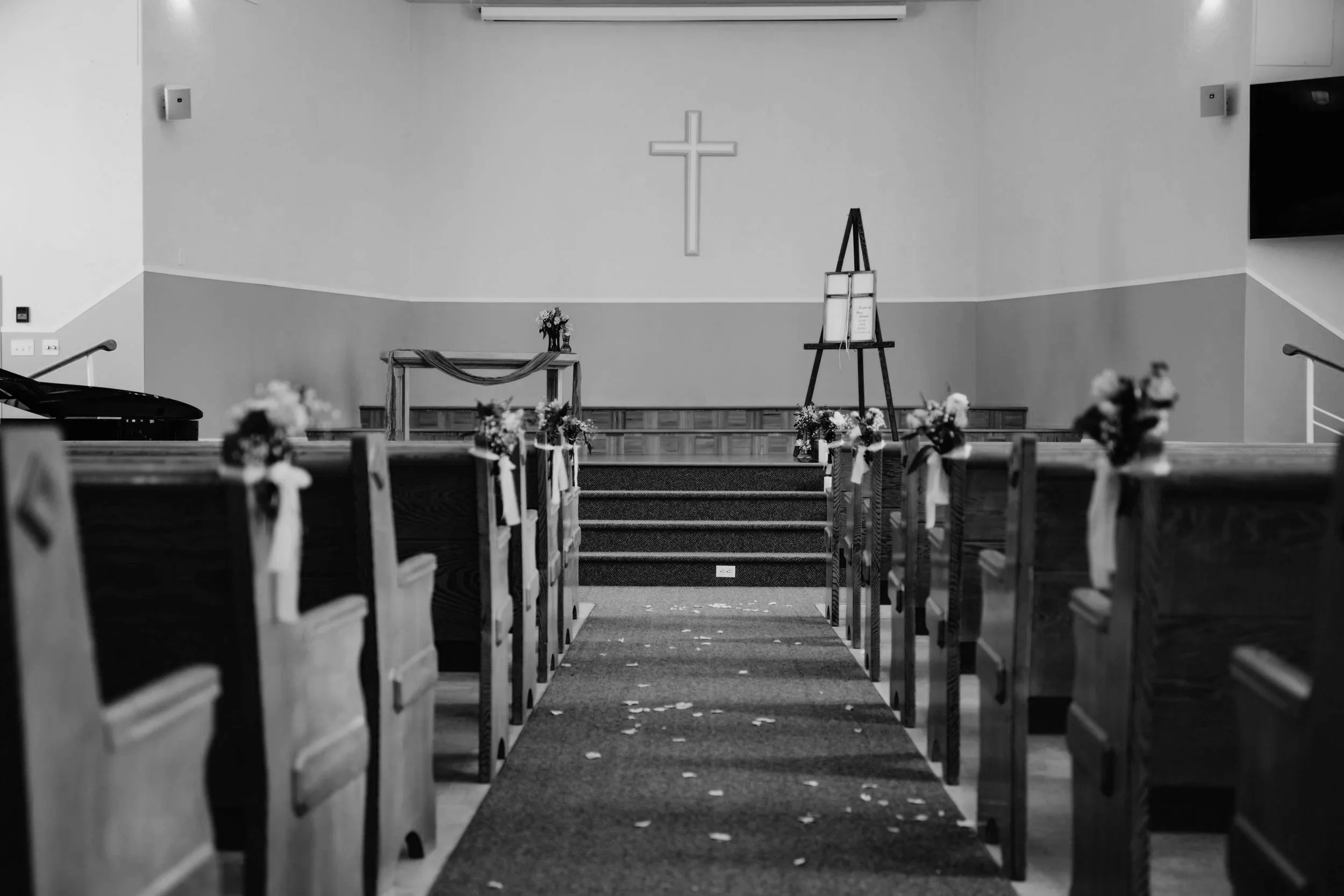 An empty church sanctuary with wooden pews decorated with flowers and ribbons, an altar with flowers, and a cross on the wall behind it. There is an easel with a framed sign beside the altar and a piano on the left side.