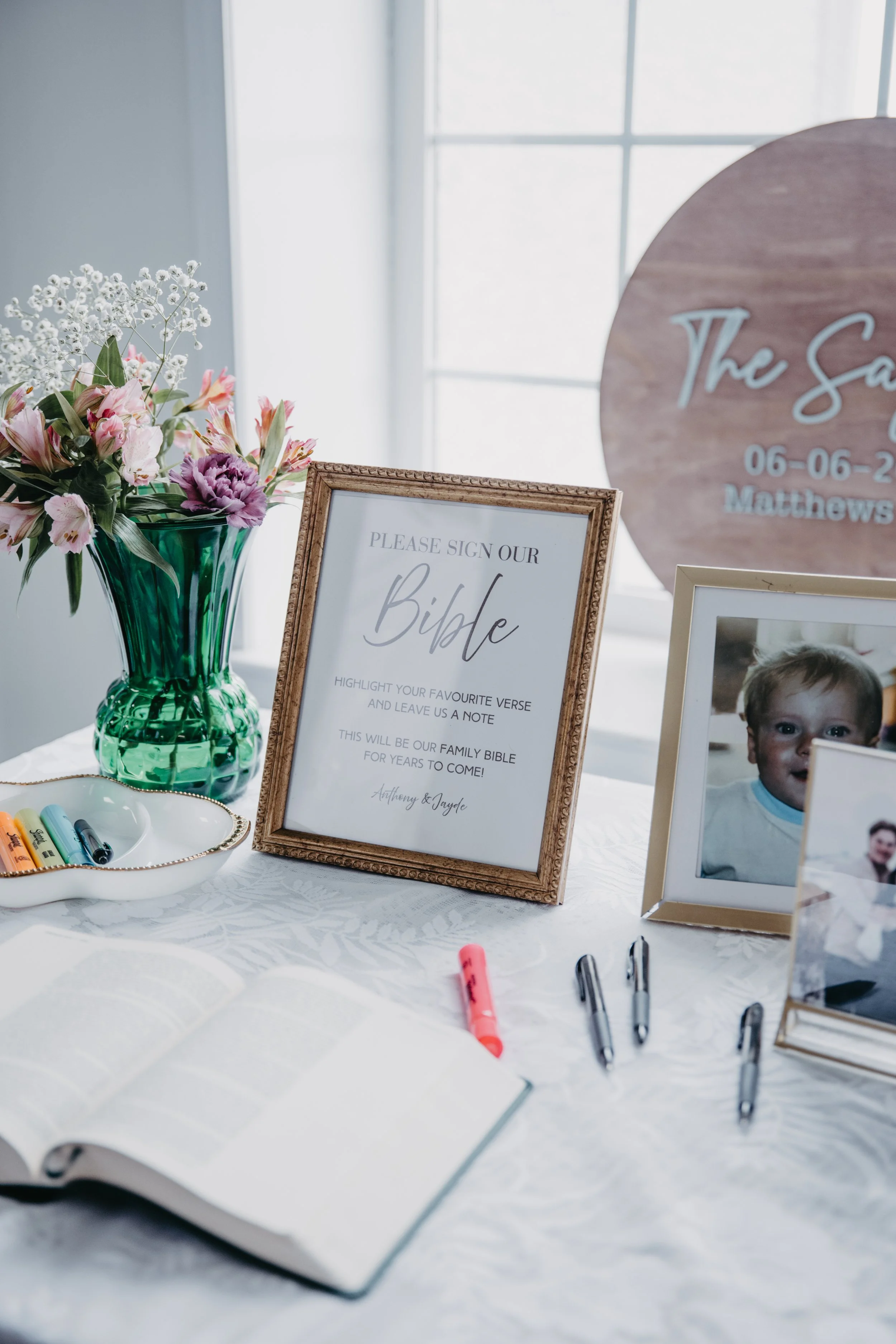 A table with a bouquet of pink and white flowers in a green glass vase, a framed sign asking guests to sign a family Bible, a framed photo of a young boy, and several pens and highlighters on a white tablecloth.