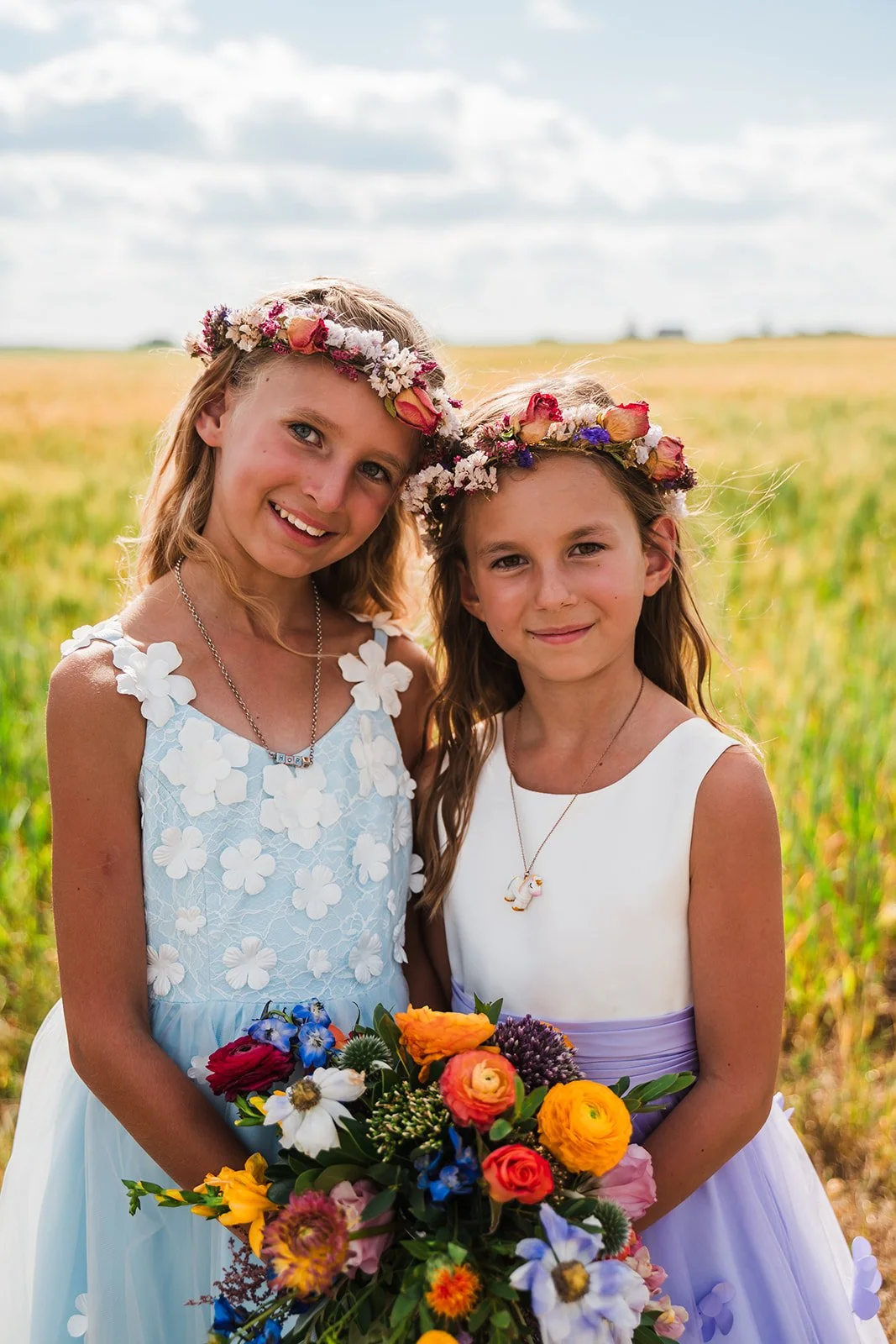 Two young girls in white dresses with floral crowns, smiling in a field, holding a bouquet of colorful flowers.