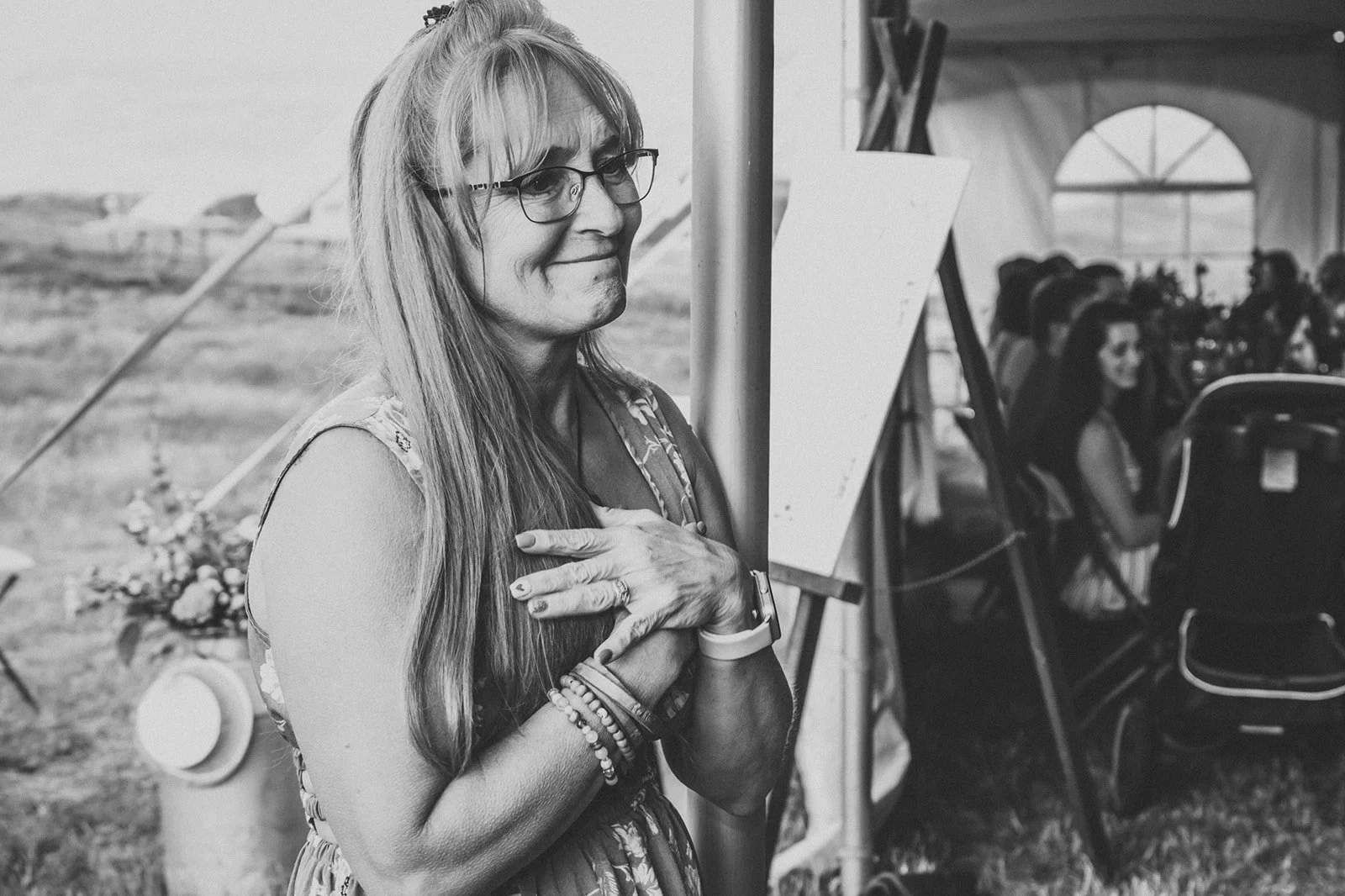 A woman with long hair and glasses, wearing a dress and multiple bracelets, stands with her hand over her heart at an outdoor event, with a tent and seated audience in the background.
