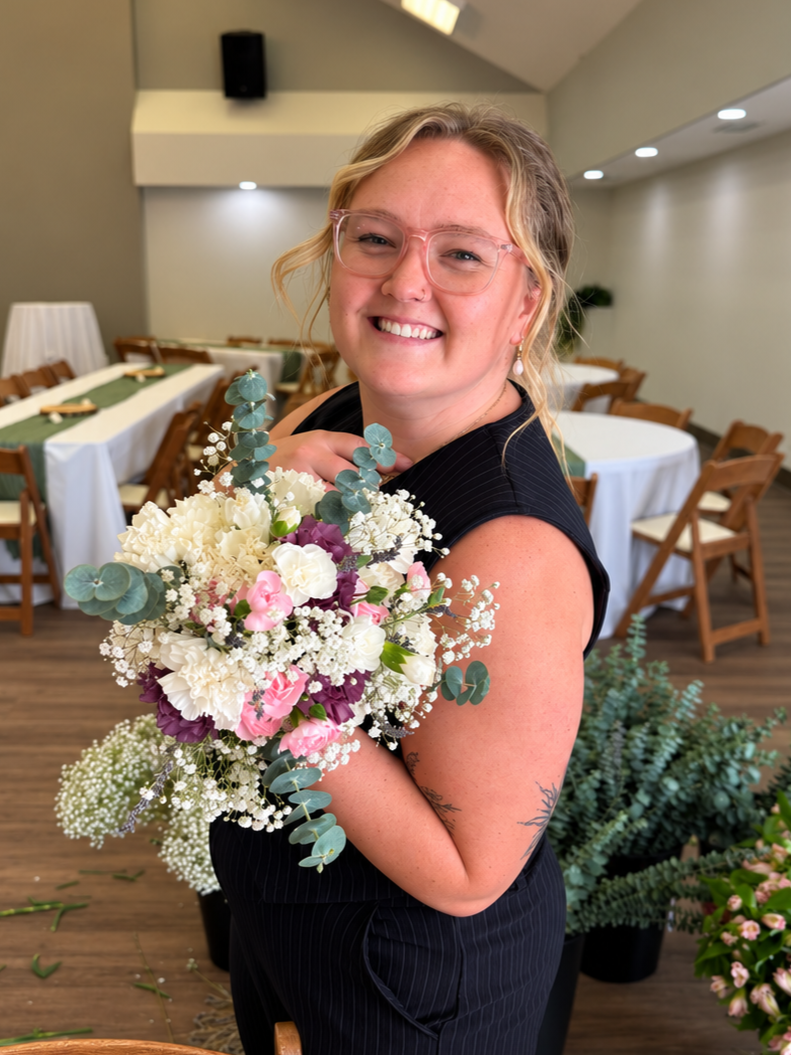 A smiling woman with blonde hair and glasses holding a bouquet of white, pink, and purple flowers in a decorated event space.
