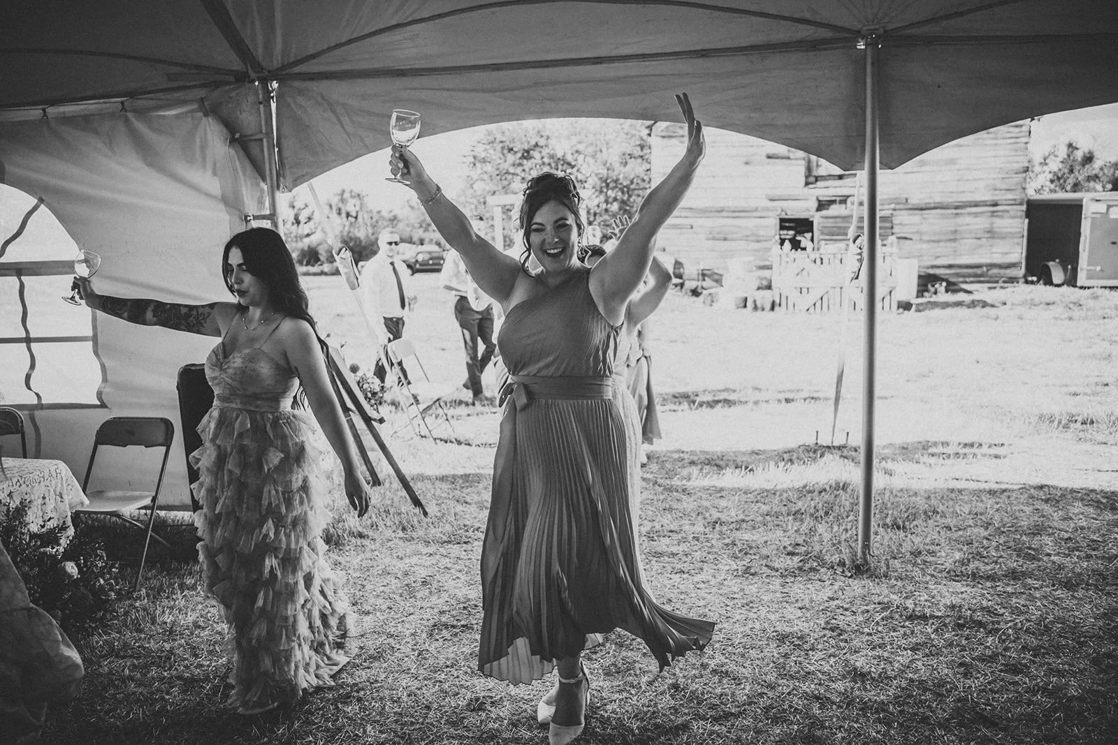 A joyful woman in a pleated dress dancing and holding a wine glass at an outdoor event, with another woman in a ruffled dress holding a wine glass nearby under a tent.