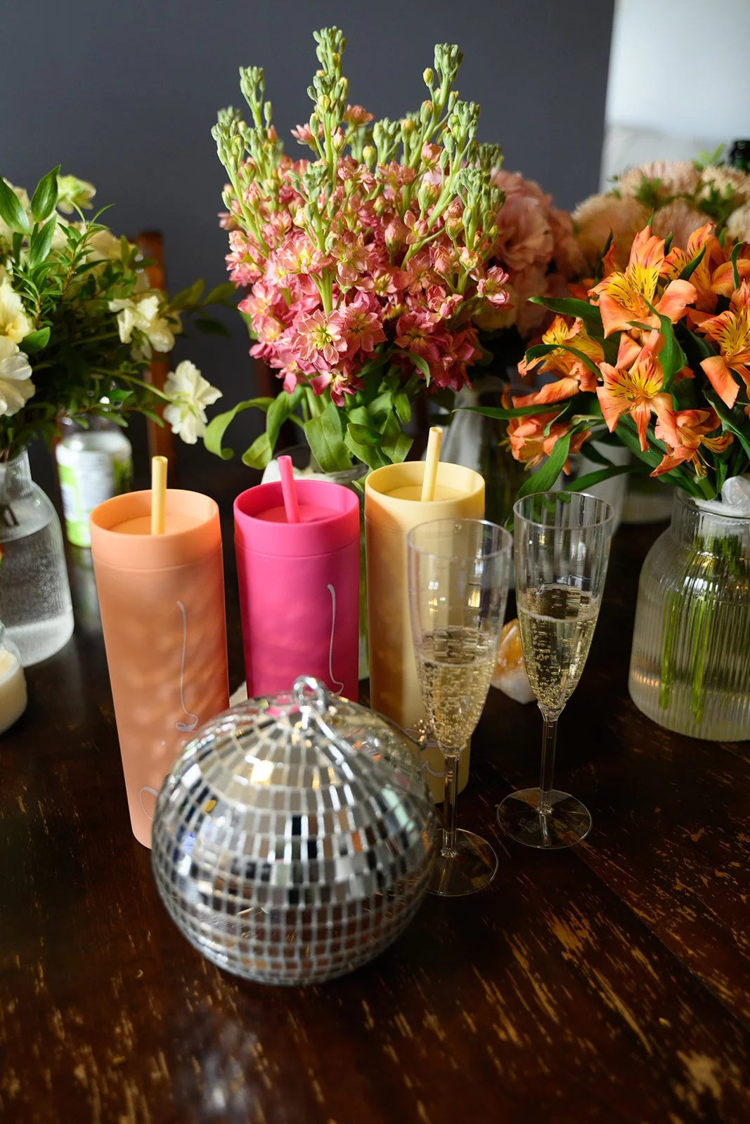 Decorative floral arrangement with pink, orange, and cream flowers, candles, and champagne glasses on a dark wooden table.