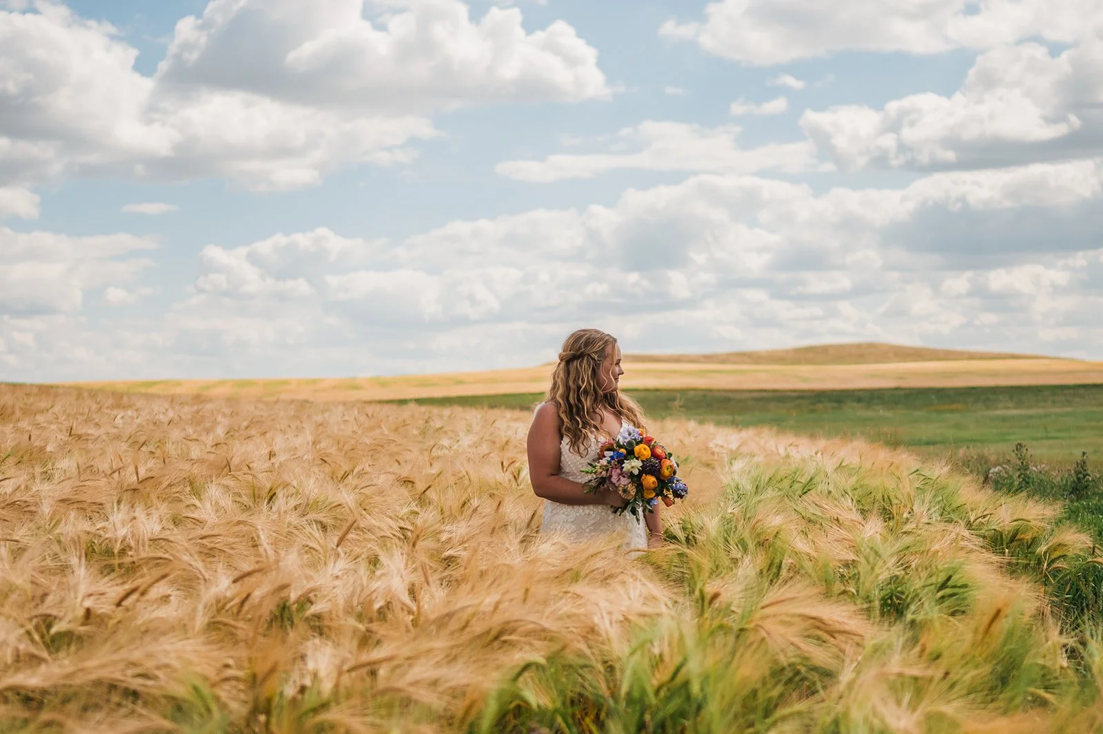 A woman wearing a white dress holding a colorful bouquet standing in a wheat field on a sunny day with a partly cloudy sky.