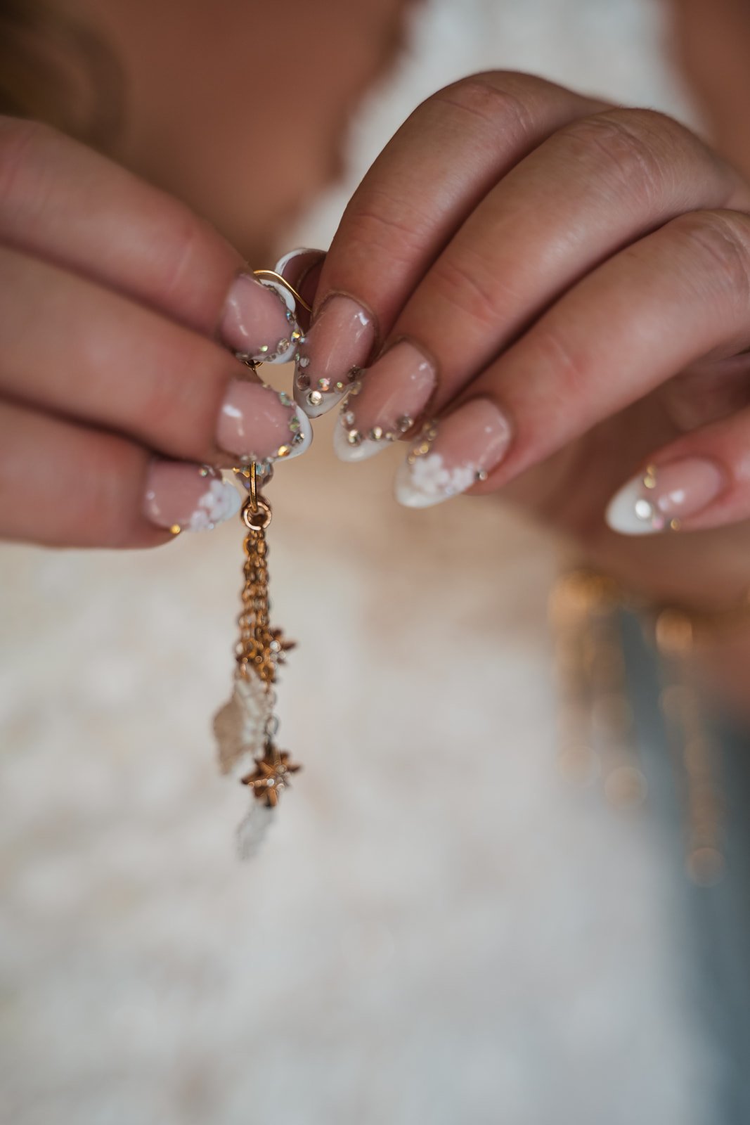 Close-up of hands holding a gold bracelet with a small charm, featuring white nail polish with decorative stones.
