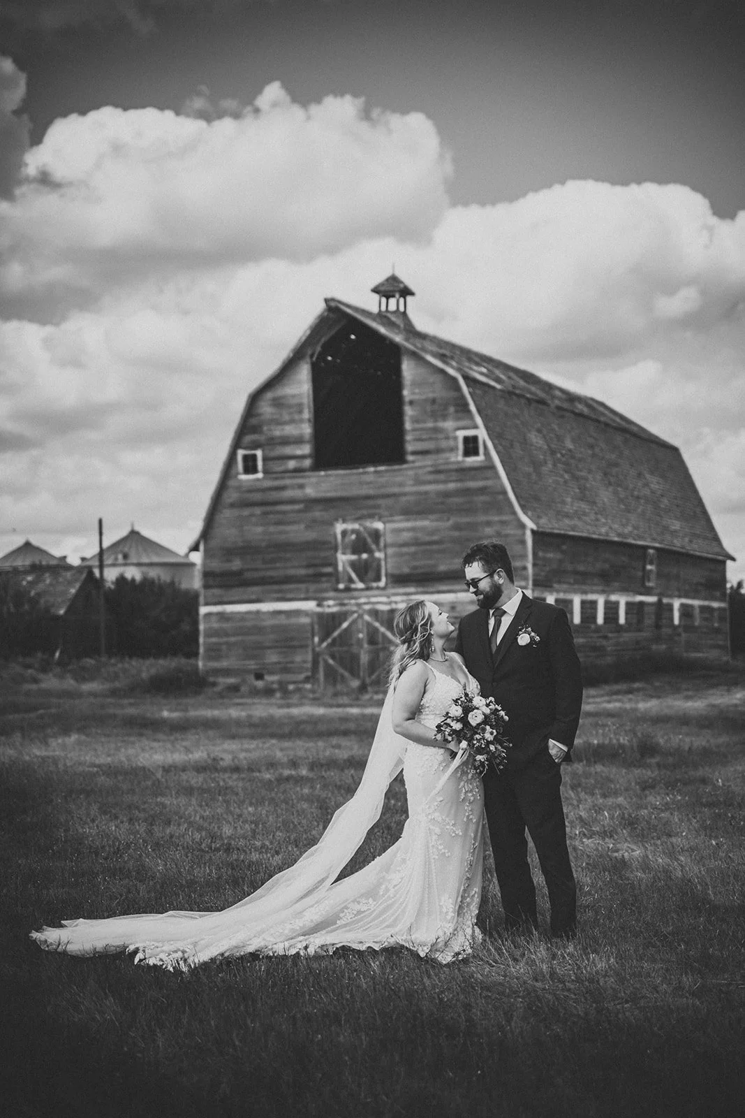A bride and groom standing on grass in front of an old barn on a cloudy day, embracing and gazing at each other, with the bride holding a bouquet of flowers.