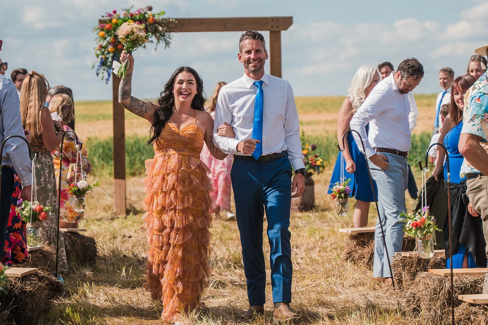 A joyful bride and groom walking down the aisle at an outdoor wedding ceremony. The bride is wearing an orange ruffled dress and holding a bouquet, while the groom is dressed in a white shirt with a blue tie and blue trousers. Guests are seated on ei
