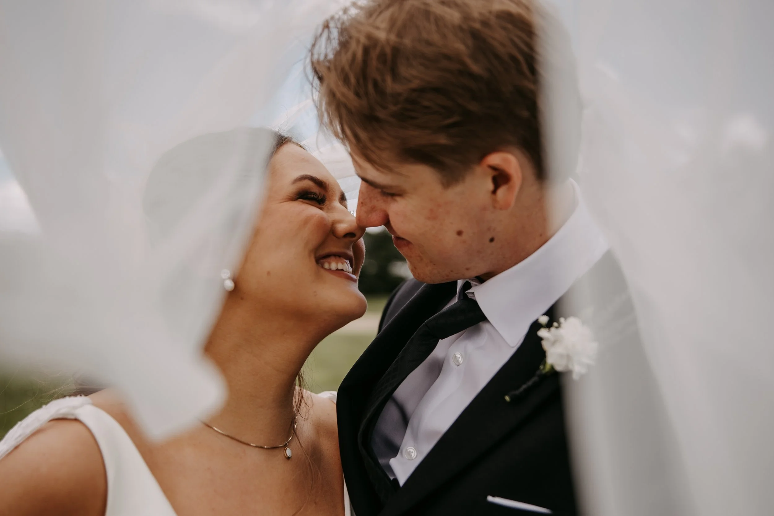 Close-up of a bride and groom smiling and touching foreheads under a white fabric, possibly a wedding ceremony.