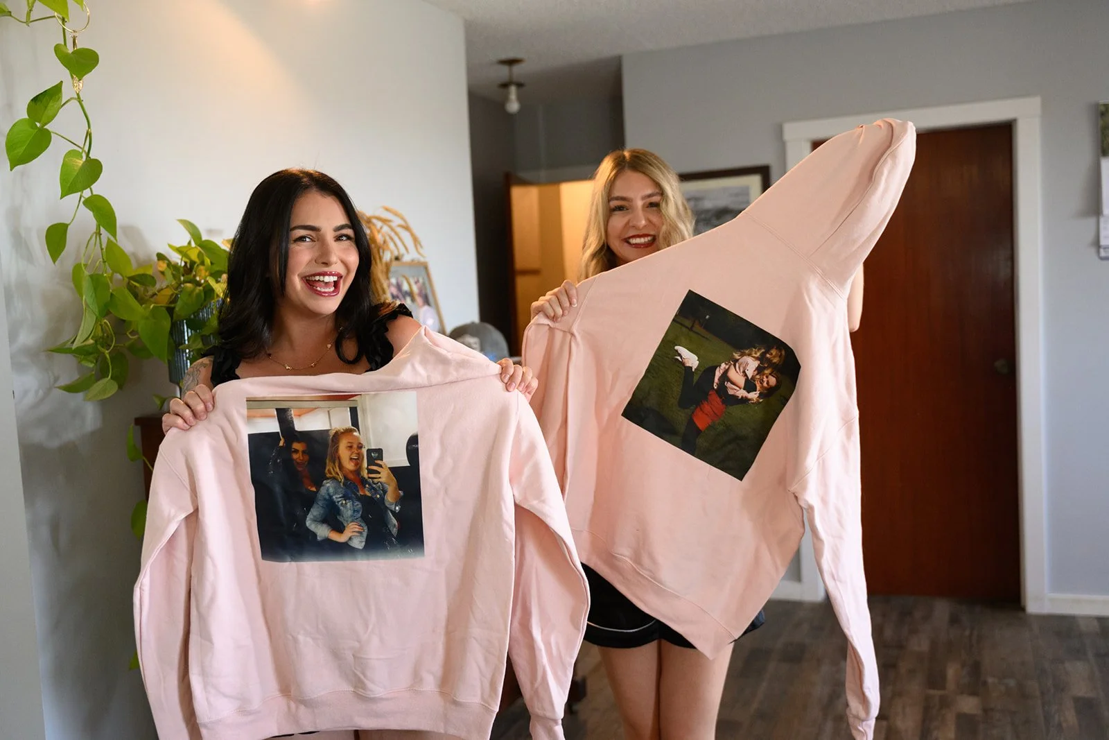 Two women smiling and holding pink sweatshirts with printed photographs of themselves on the front. The women are in a living room with a plant, a framed photo, and a doorway in the background.