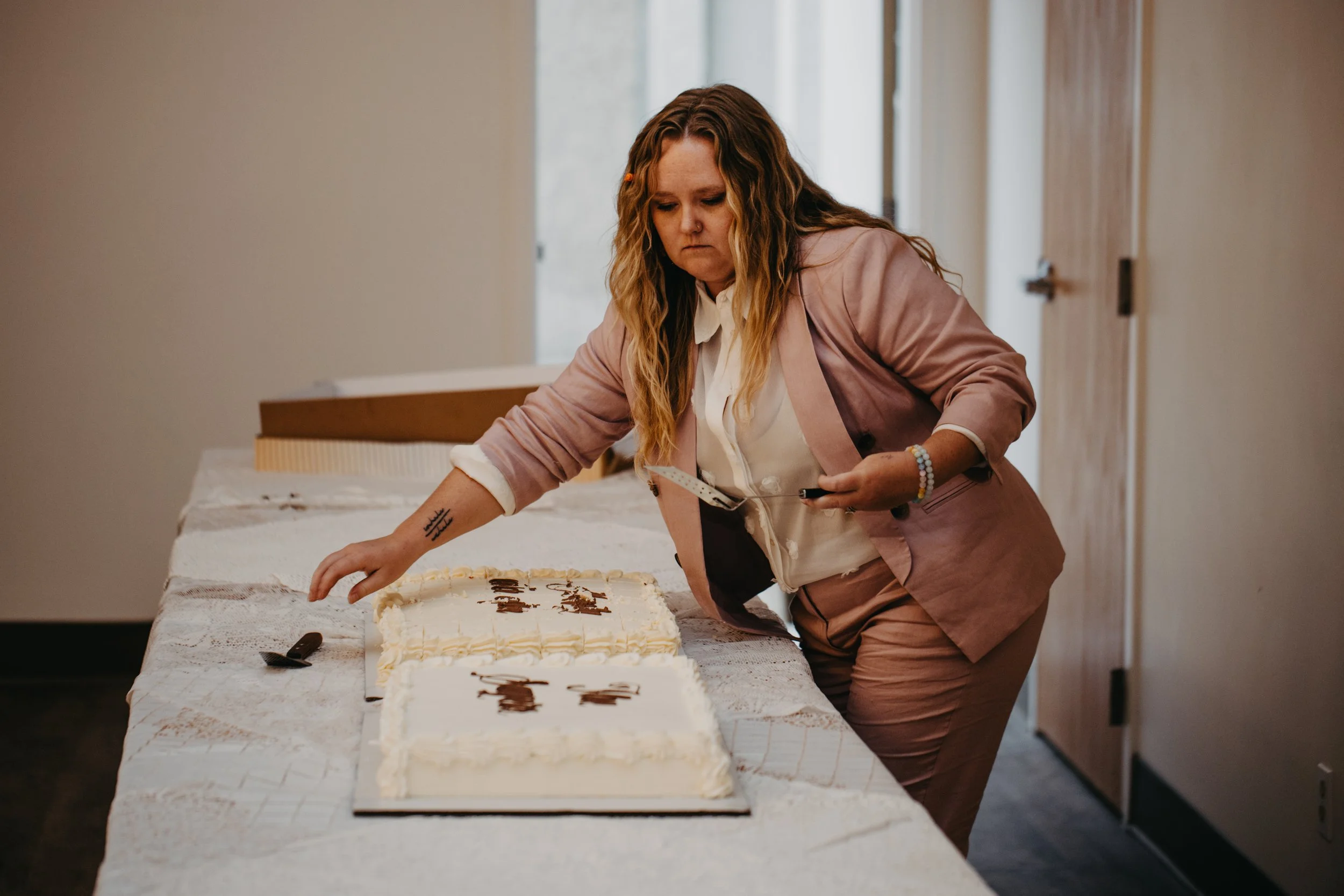 A woman with wavy, reddish-brown hair wearing a pink blazer and white shirt, leaning over a table with a large white cake, writing or decorating on it with a small tool.