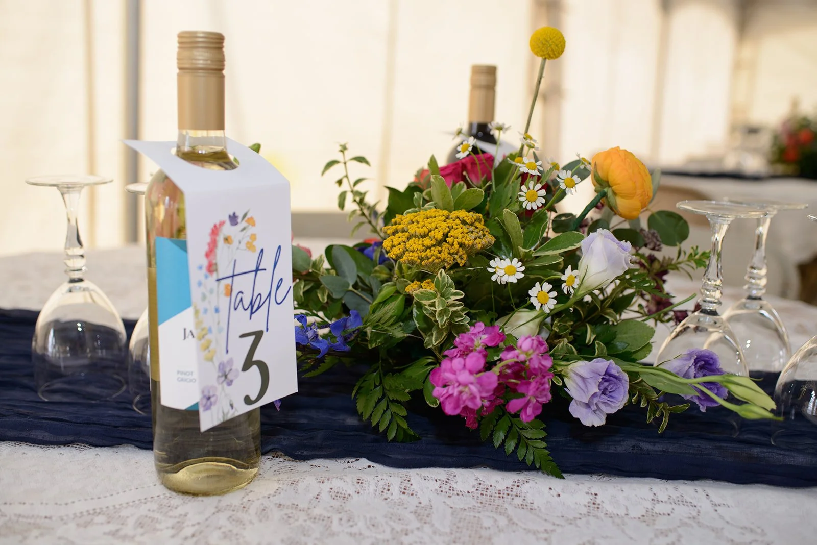 A bottle of white wine with a table number tag marked 'Table 3' placed in front of a floral centerpieces with various colorful flowers, surrounded by upside-down wine glasses on a table with a lace tablecloth.